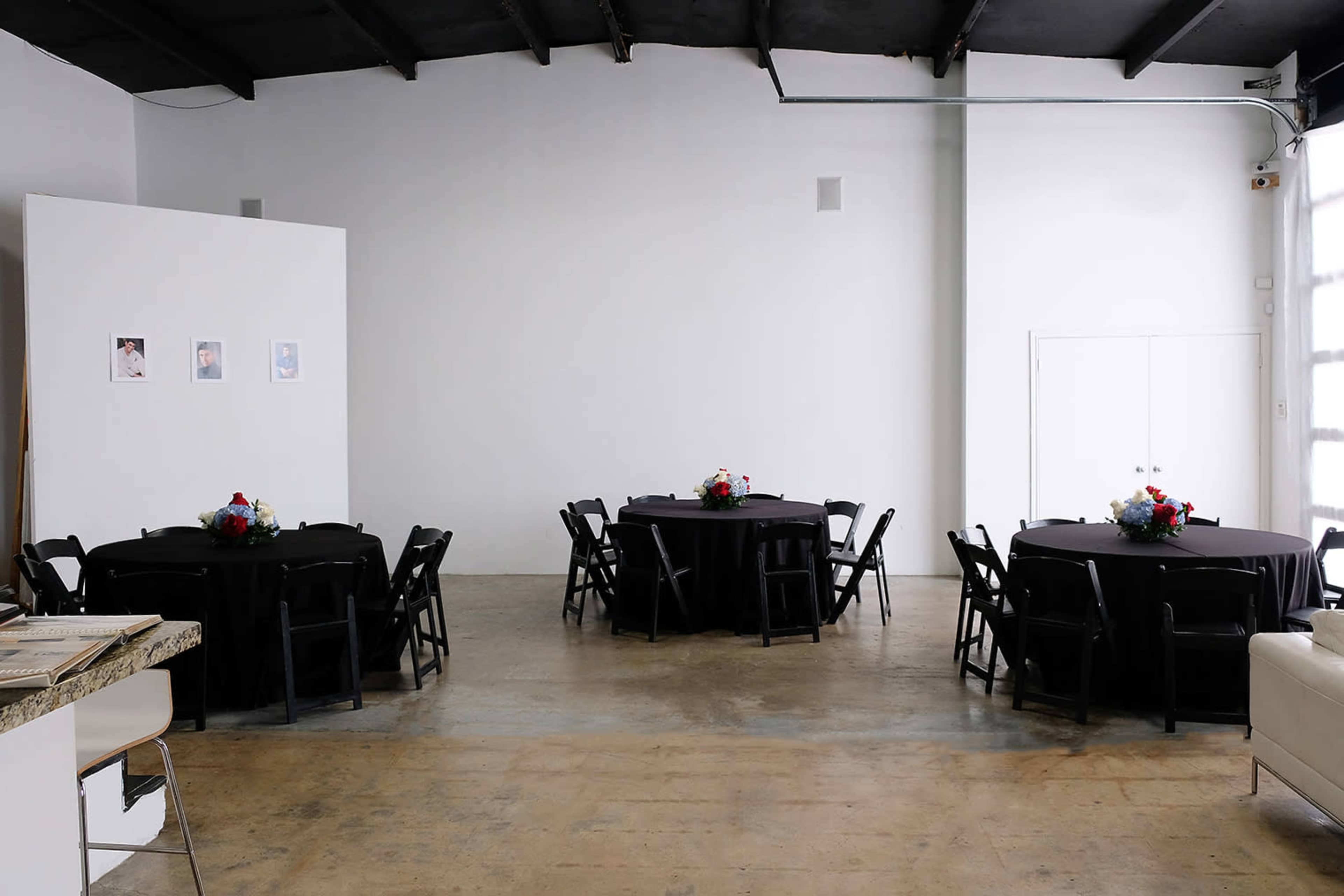 A spacious event venue with two round tables covered in black cloth, each adorned with floral centerpieces, set against a plain white wall.