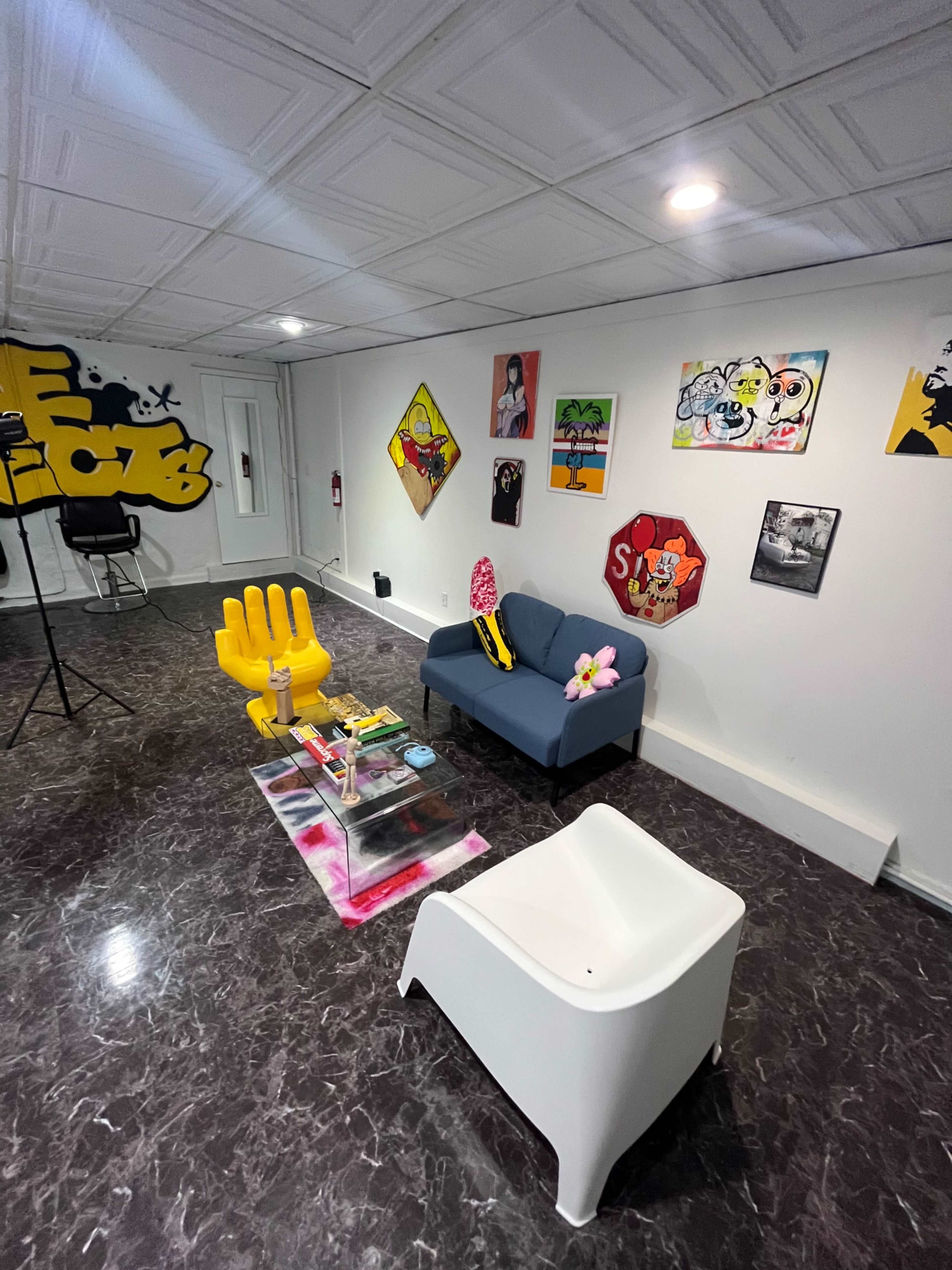 A modern room featuring a gray sofa, colorful artwork on the walls, a yellow hand-shaped chair, and a white chair on a glossy dark floor.