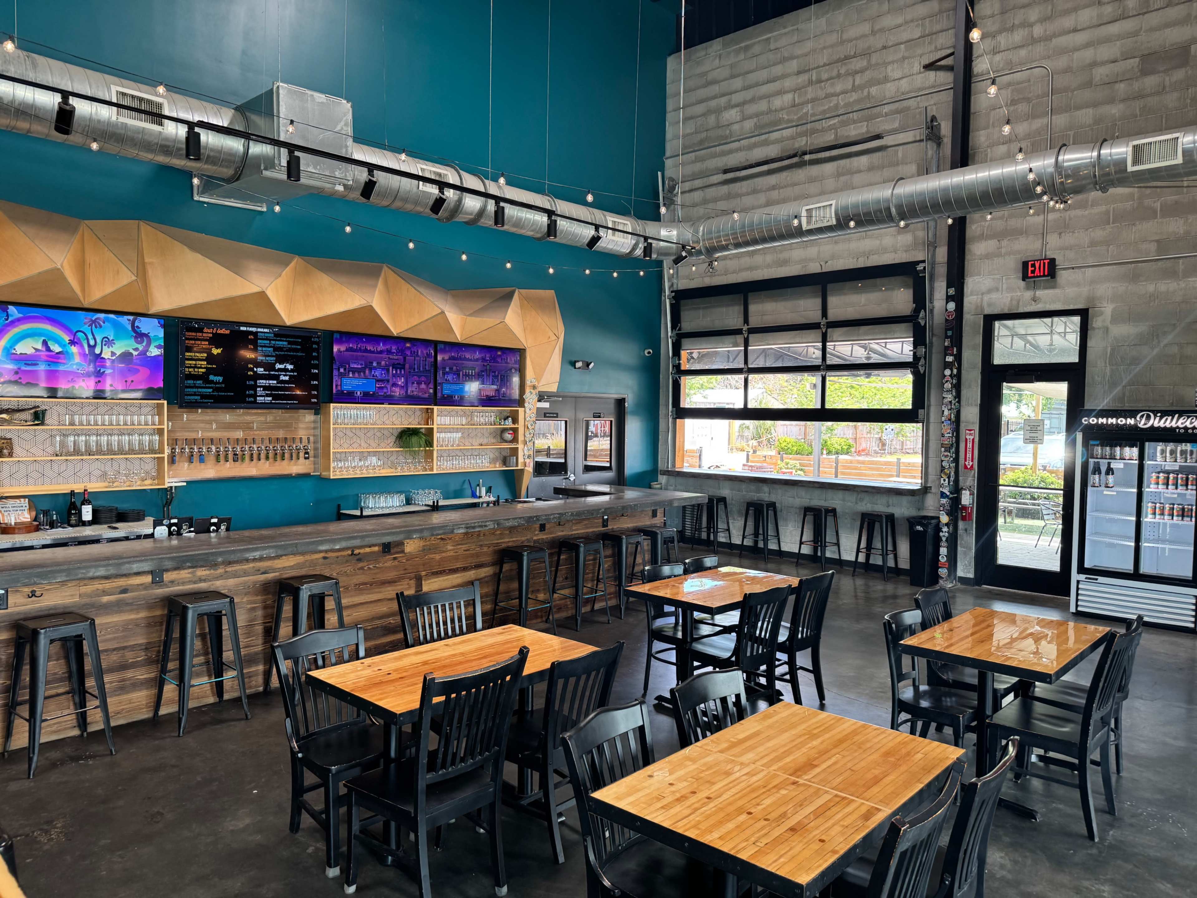 The image shows a modern brewpub interior with wooden tables, a bar area, and a large backlit menu display on a colorful wall.