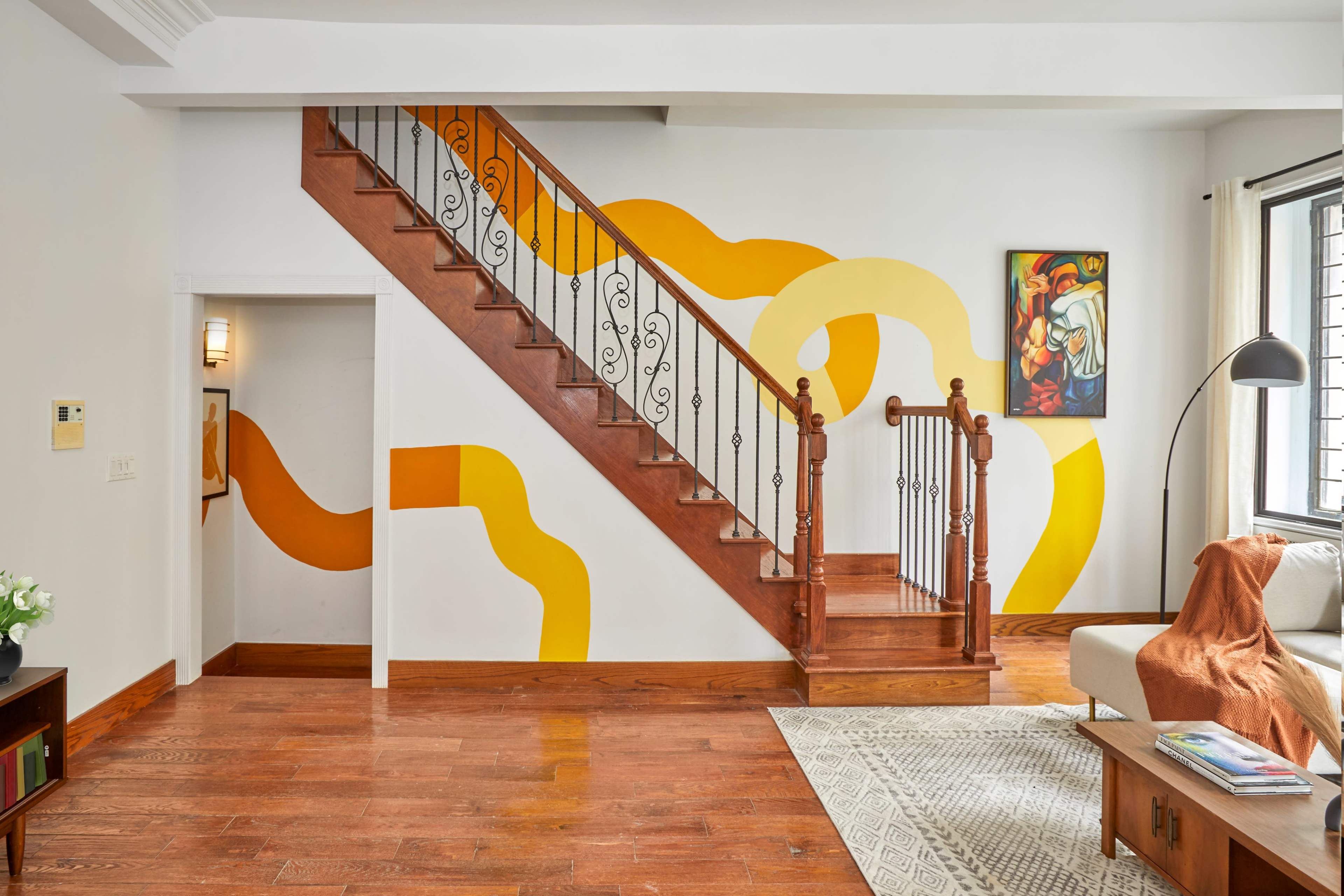 A wooden staircase with decorative railings ascends against a wall featuring abstract yellow and orange patterns, adjacent to a sitting area with a lamp and an art piece.