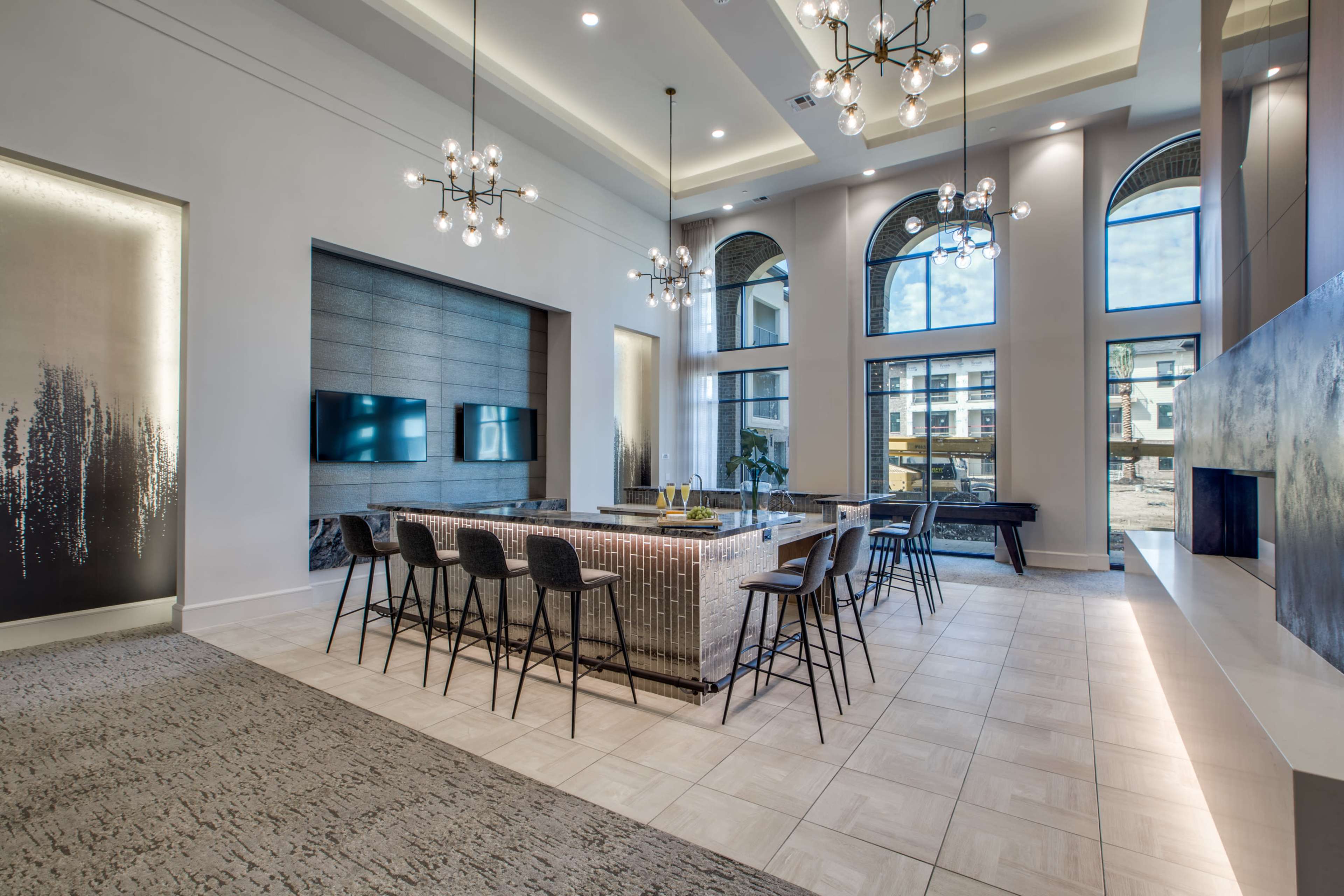 The image shows a modern interior space featuring a large communal area with high ceilings, a stylish kitchen island, and multiple seating arrangements under decorative light fixtures.