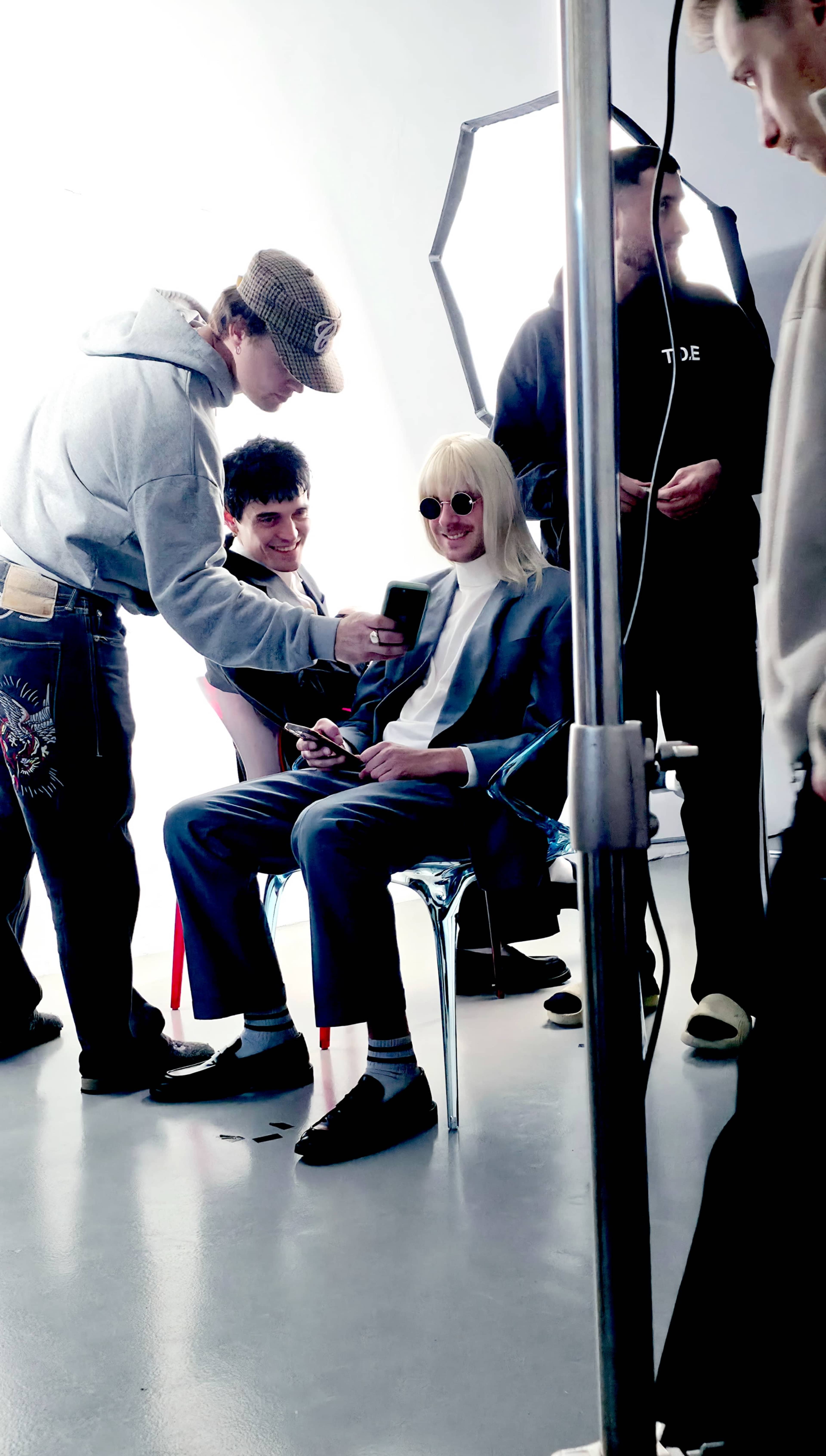 A group of people is gathered in a studio, with one person seated in a chair wearing sunglasses and a wig while others assist with a phone and equipment.