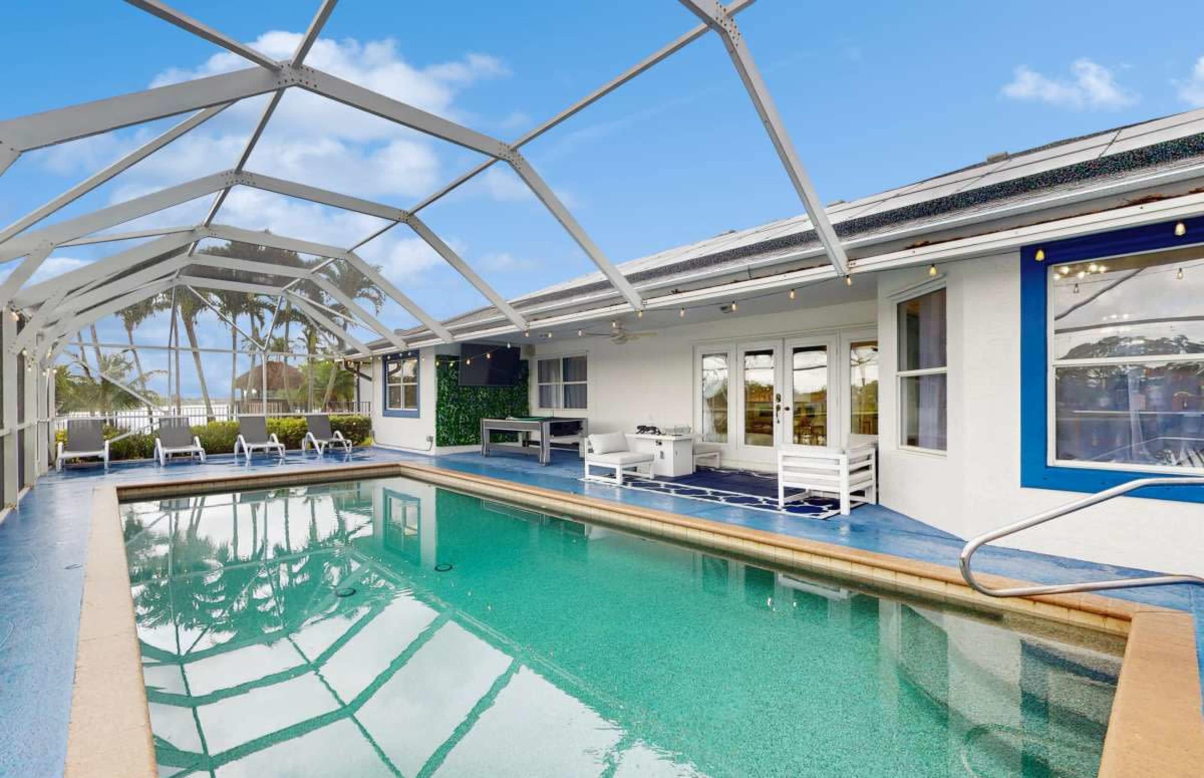 The image shows a clear swimming pool with a screened enclosure and lounge chairs beside it, located next to a bright, modern house.