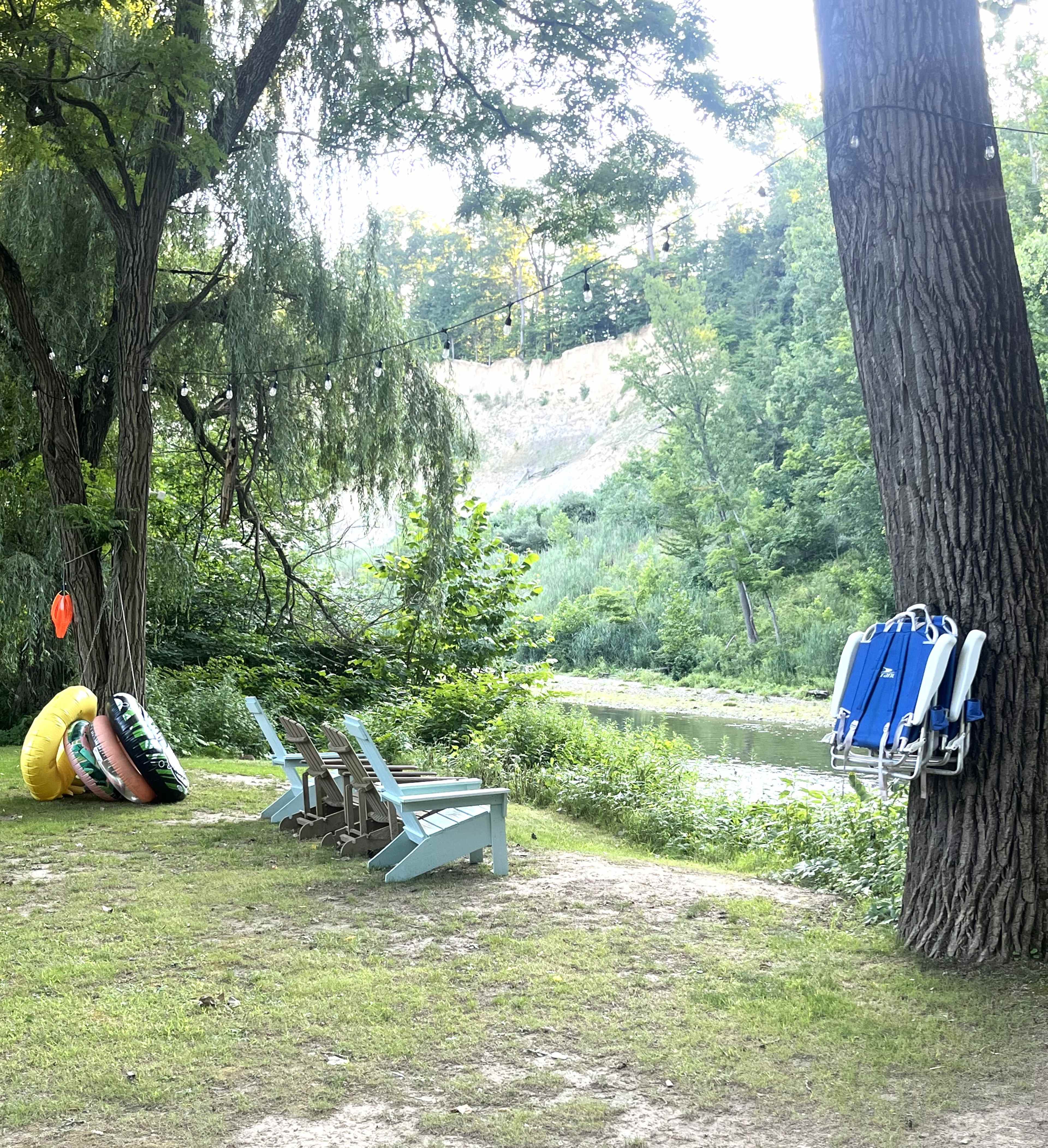A grassy area beside a river features a set of adirondack chairs and colorful inflatable flotation devices hanging from a tree.