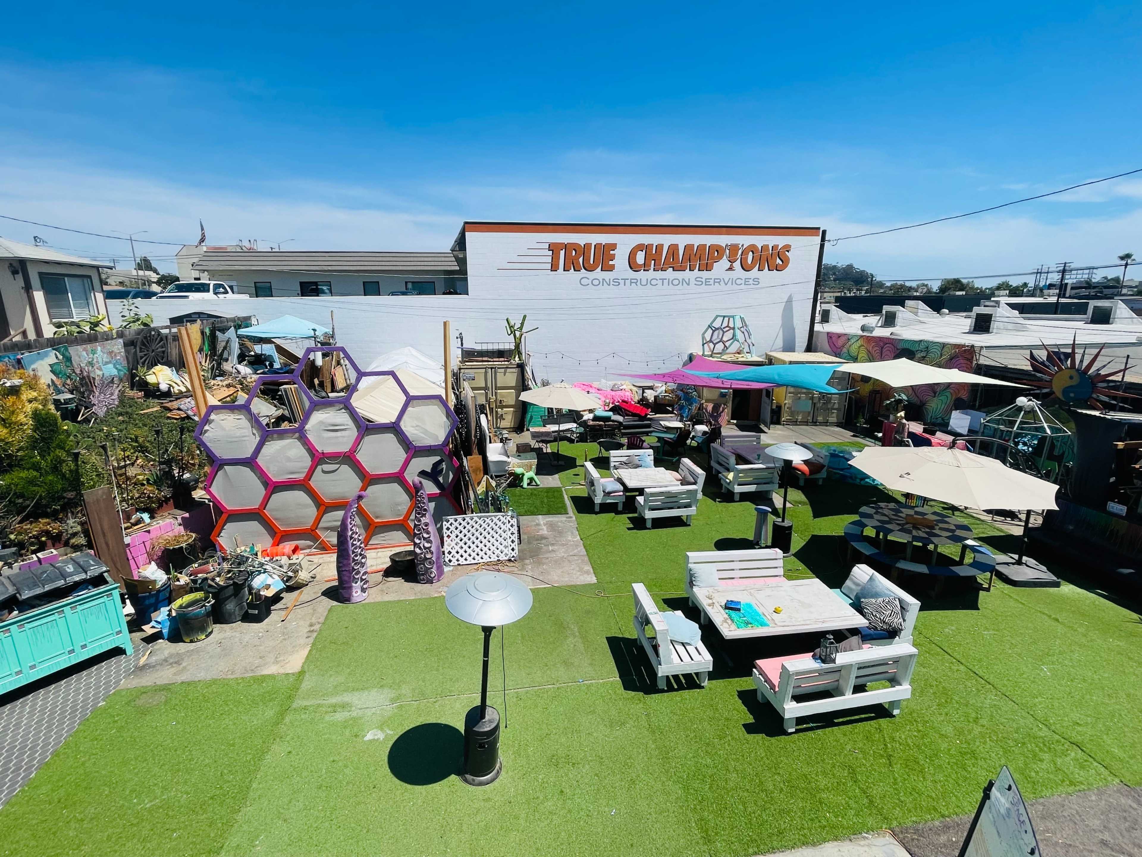 A colorful outdoor space with hexagonal decorations, seating areas, umbrellas, and various plants against a backdrop of a white building displaying the name "True Champions Construction Services."
