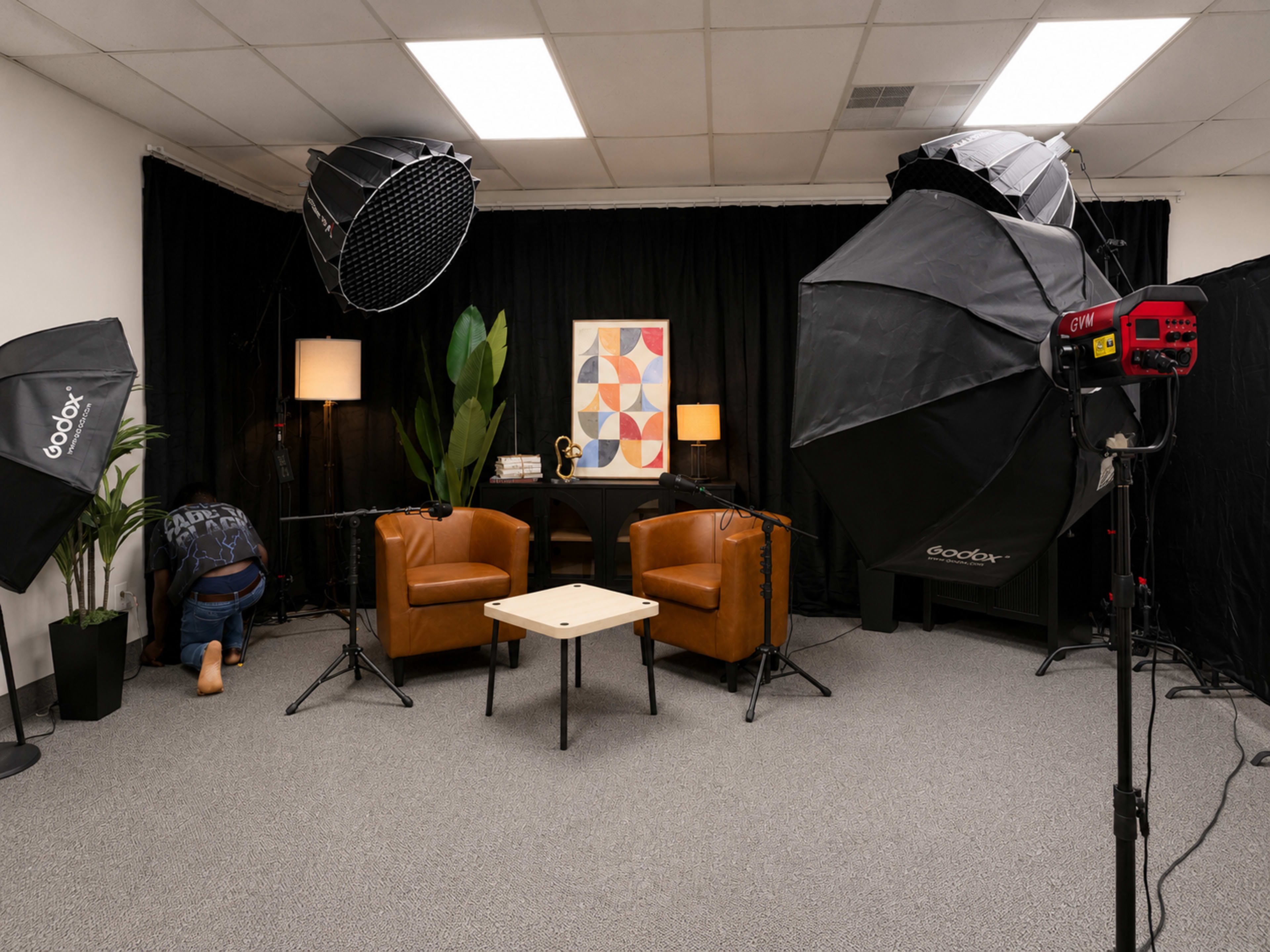 The image shows a studio setup with two brown chairs, a small table, and professional lighting equipment, all arranged against a black curtain backdrop.