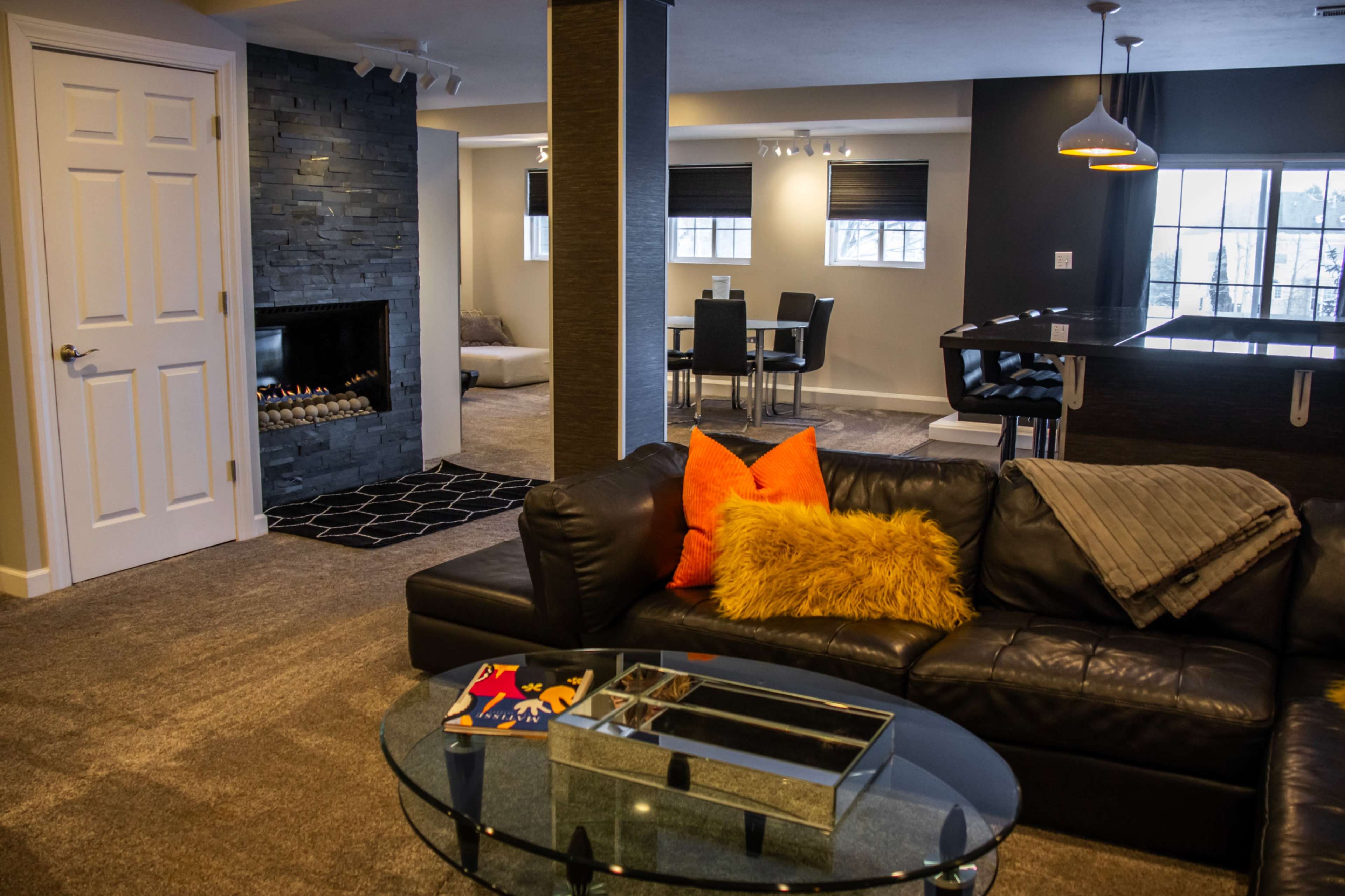 A modern living space features a black leather sofa with colorful cushions, a glass coffee table, a stone fireplace, and a dining area in the background.