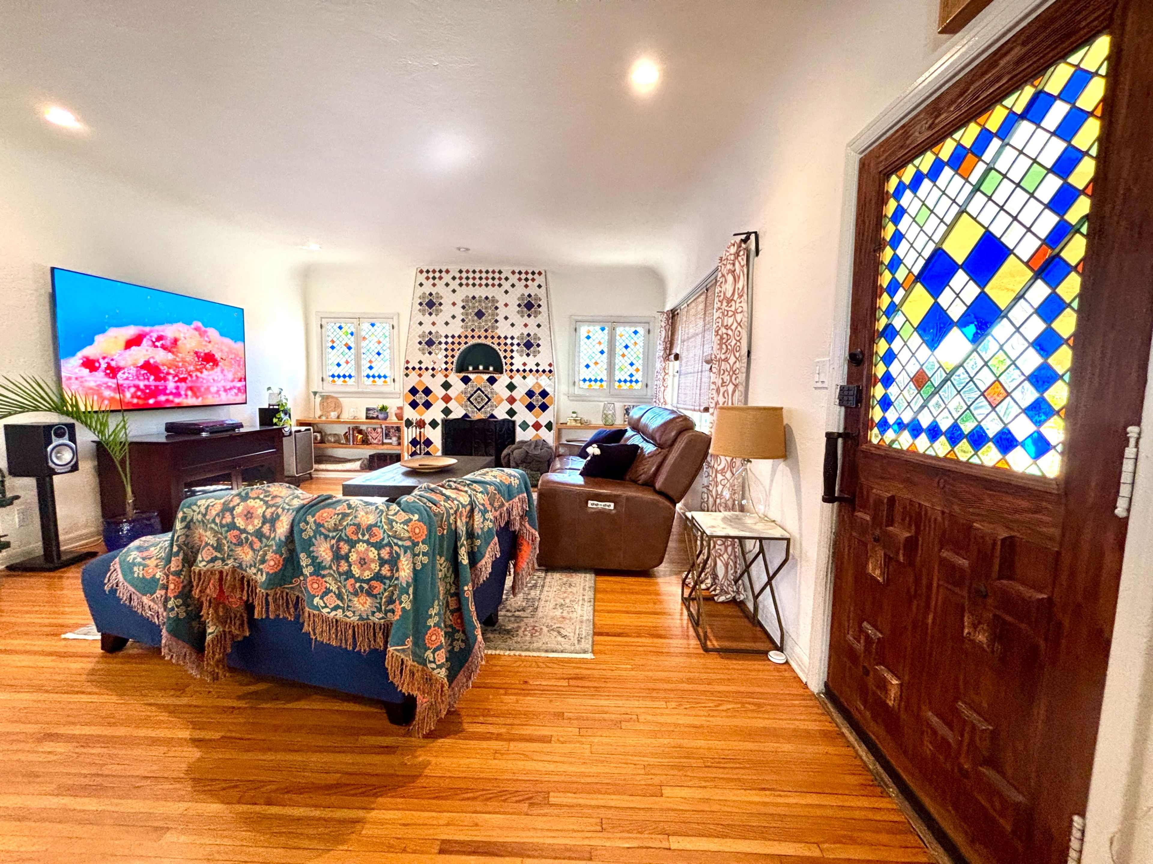 The image shows a living room with a large television, colorful stained glass windows, and a combination of seating arrangements, including a sofa and an armchair.