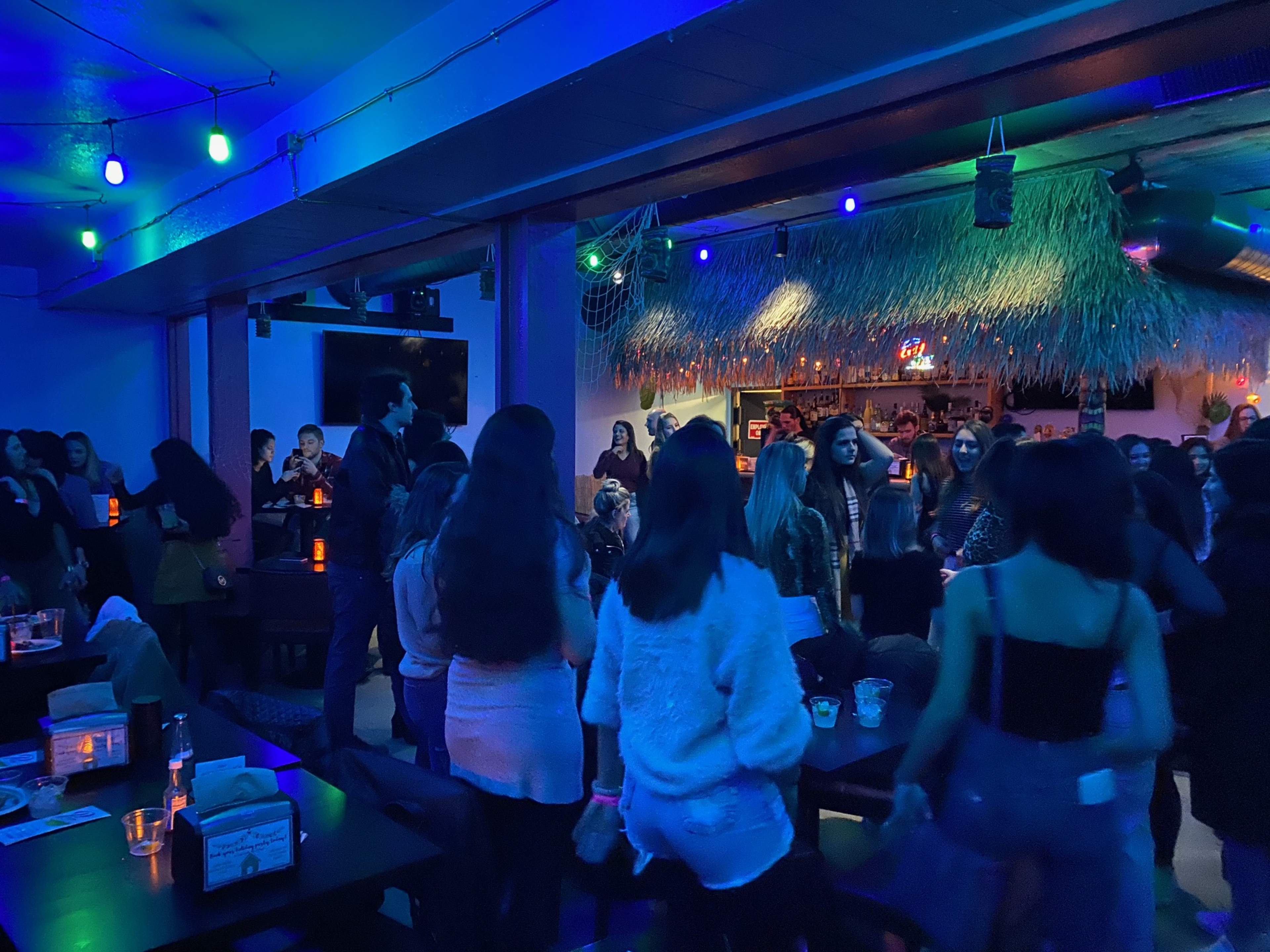 A lively crowd gathers in a dimly lit bar adorned with colorful lights and a thatched roof.