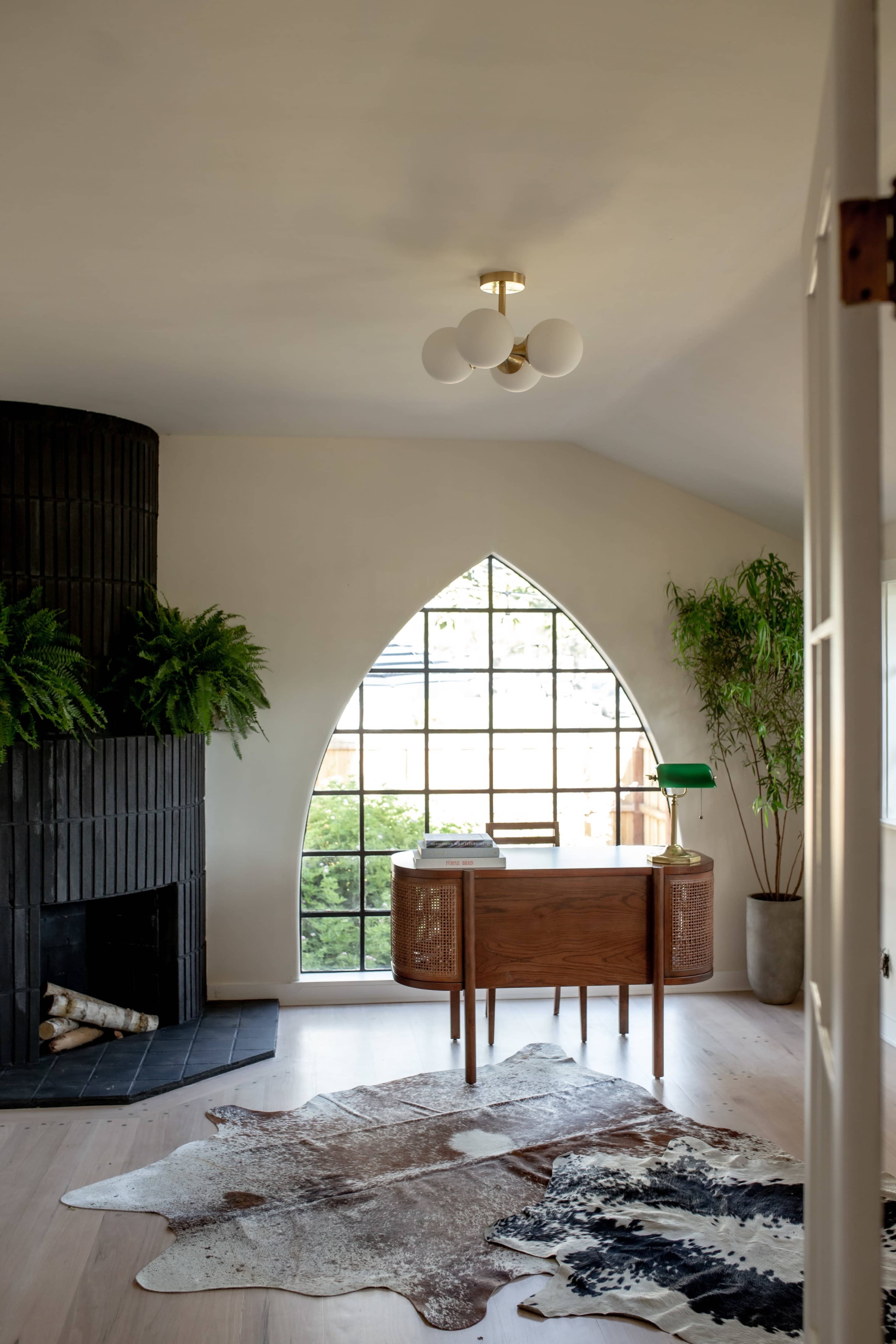 A modern interior with a wooden desk positioned beneath an arched window, a black fireplace, and greenery in decorative pots.