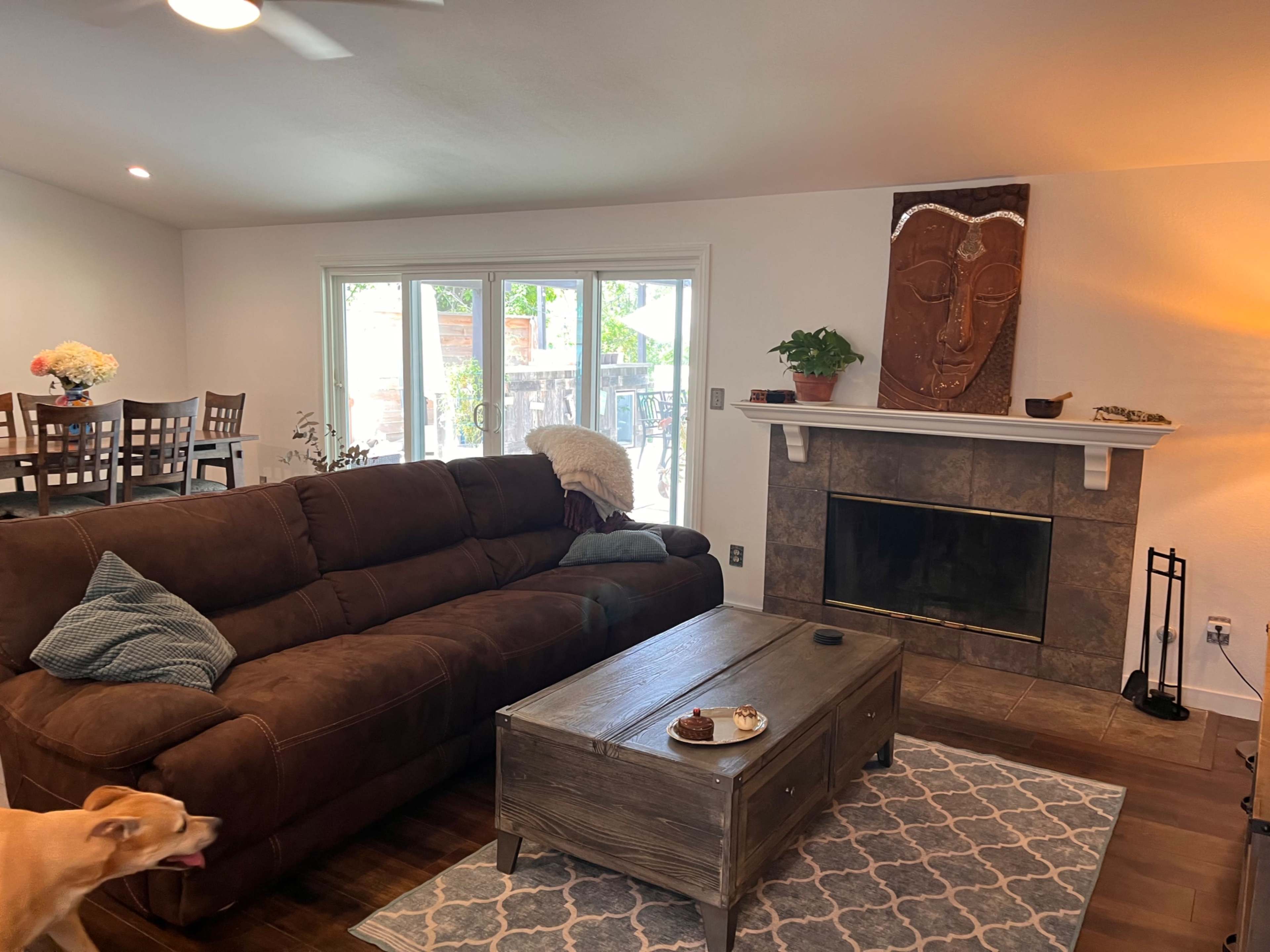 A living room featuring a brown sectional sofa, a wooden coffee table, and a carved wall hanging above a fireplace, with a dining area visible in the background.