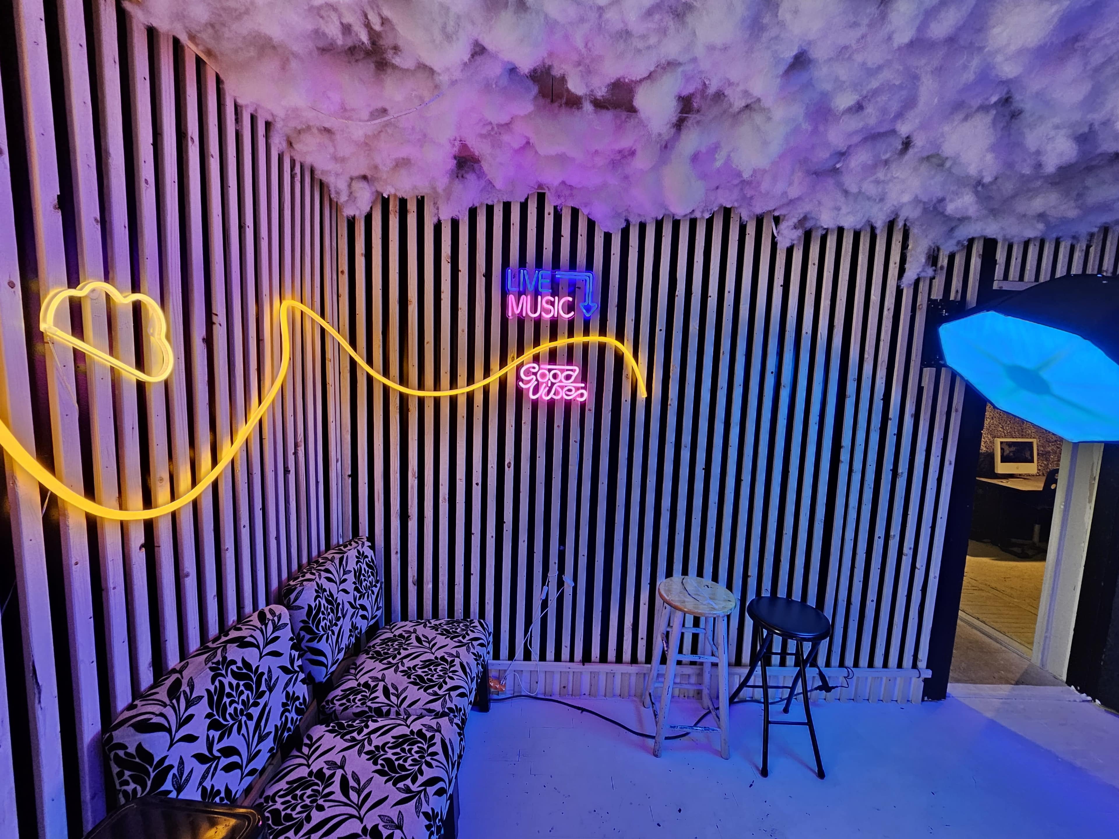 The image shows a room with a black and white striped wall, a neon sign reading "LIVE MUSIC," a cloud-shaped neon light, and modern furniture including a patterned couch and stools, surrounded by a ceiling decorated with white fluffy material.