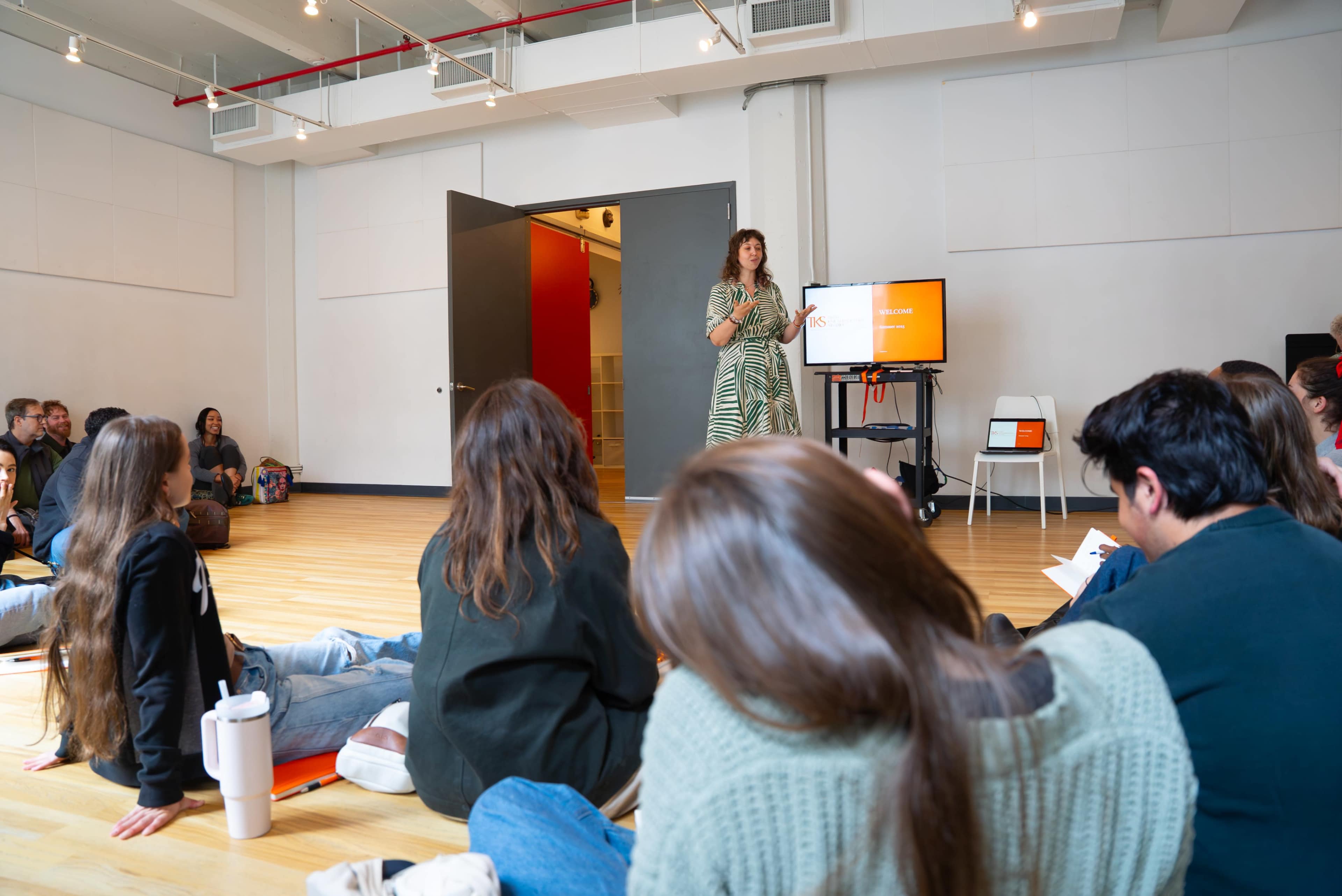 A woman stands in front of an audience seated on the floor in a bright, open room, delivering a presentation.
