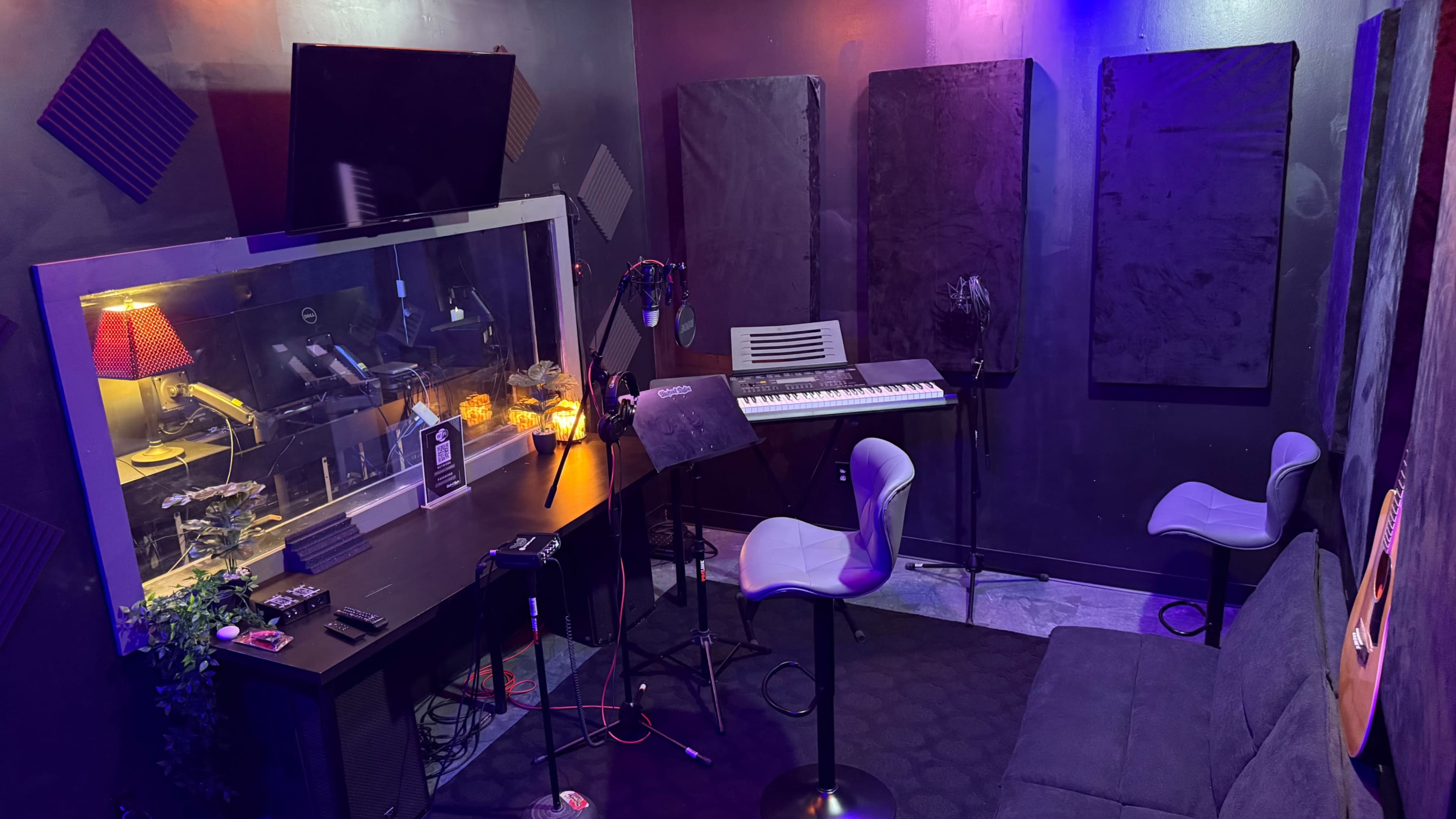 The image shows a small recording studio with soundproofing panels, a keyboard, a microphone setup, and ambient lighting.