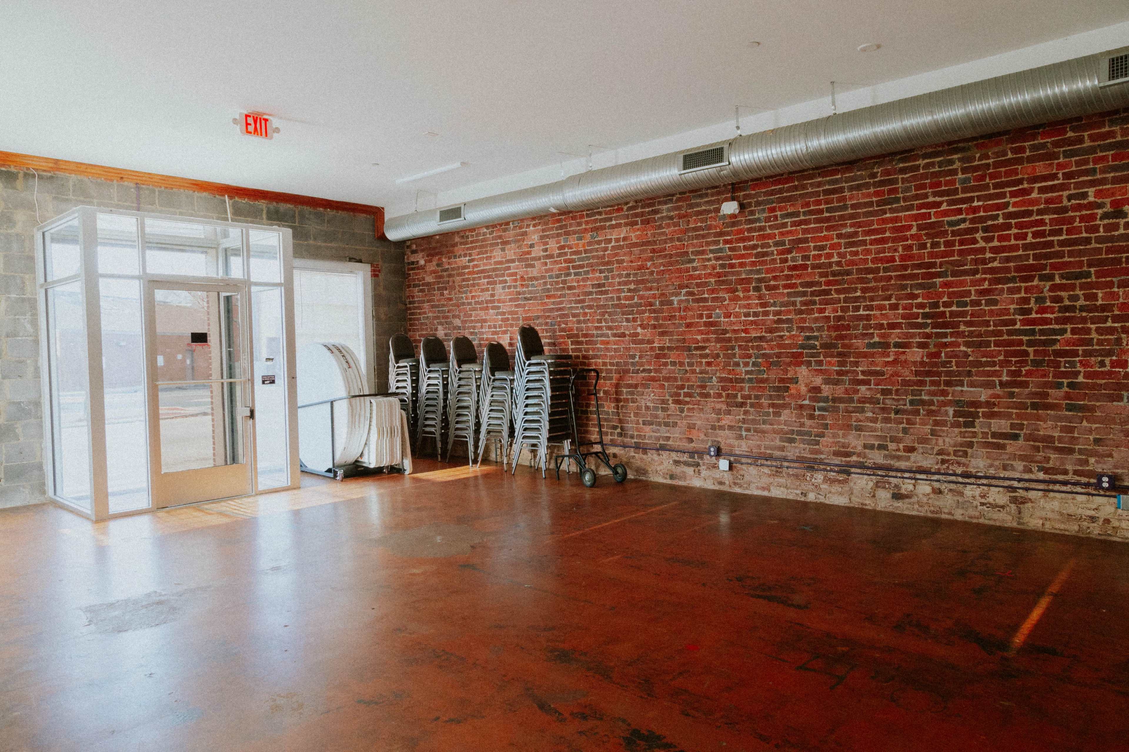 The image shows an empty room with a brick wall, polished concrete floor, and several metal folding chairs stacked against a wall.