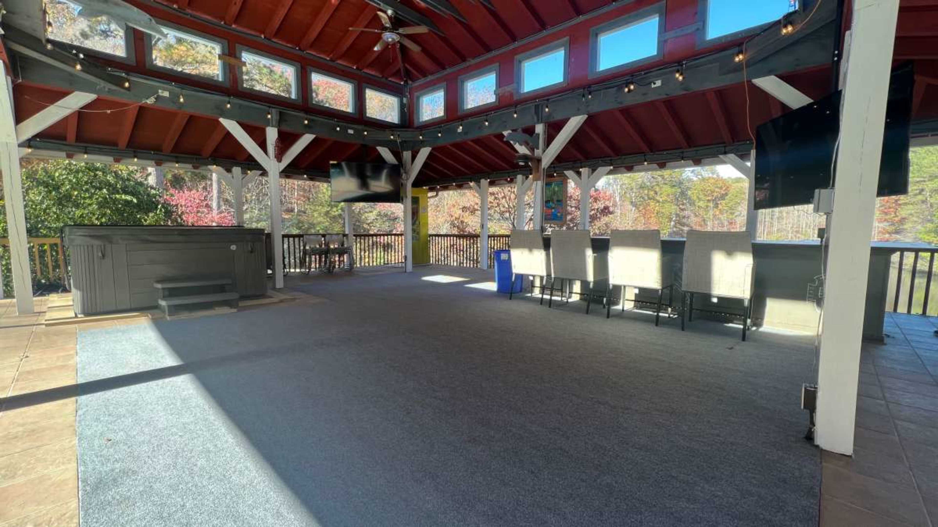 The image shows an open pavilion with a hot tub, seating area, and large windows, surrounded by a wooded landscape.