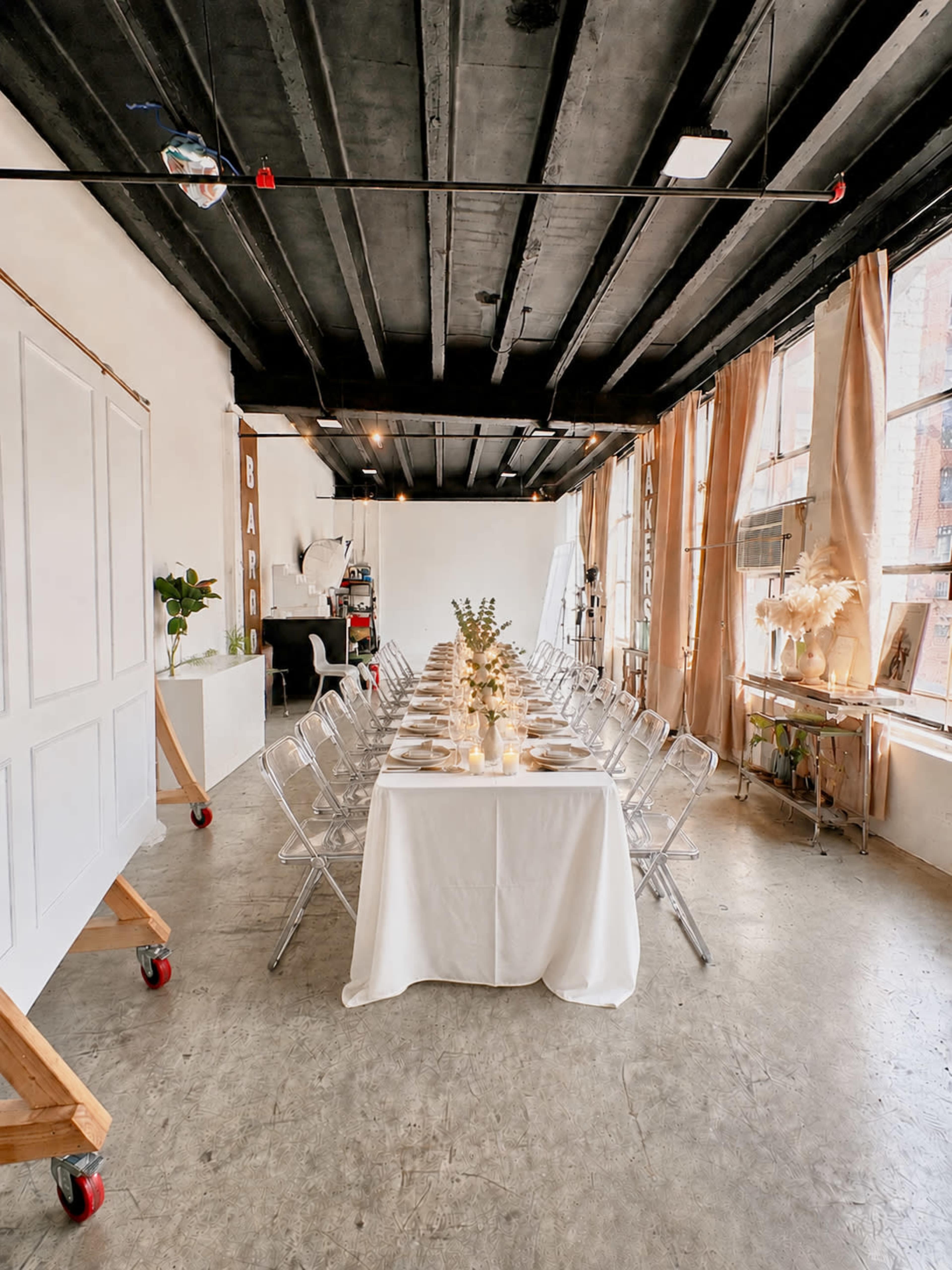 The image shows a long dining table set with a white tablecloth, surrounded by transparent chairs, in a modern industrial space with large windows and minimal decor.