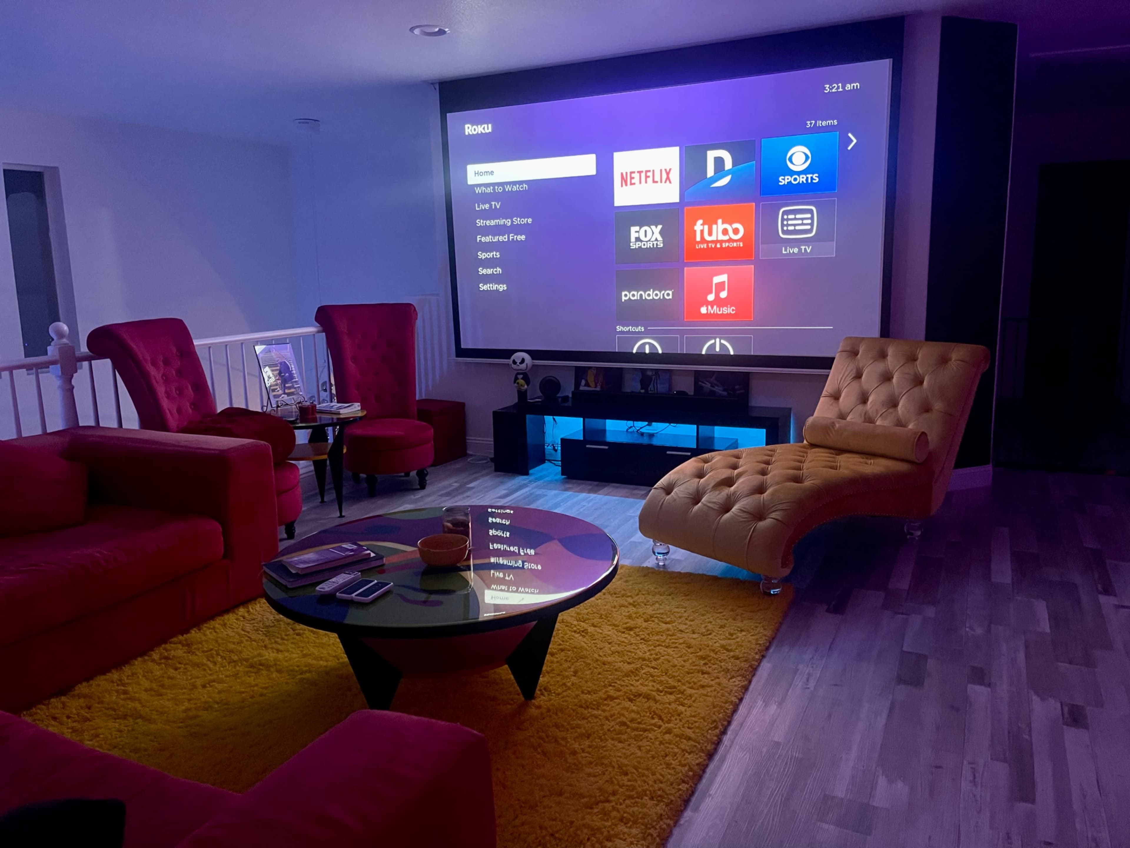 A modern living room with a large projector screen displaying various streaming services, surrounded by red and yellow furniture on a wooden floor.