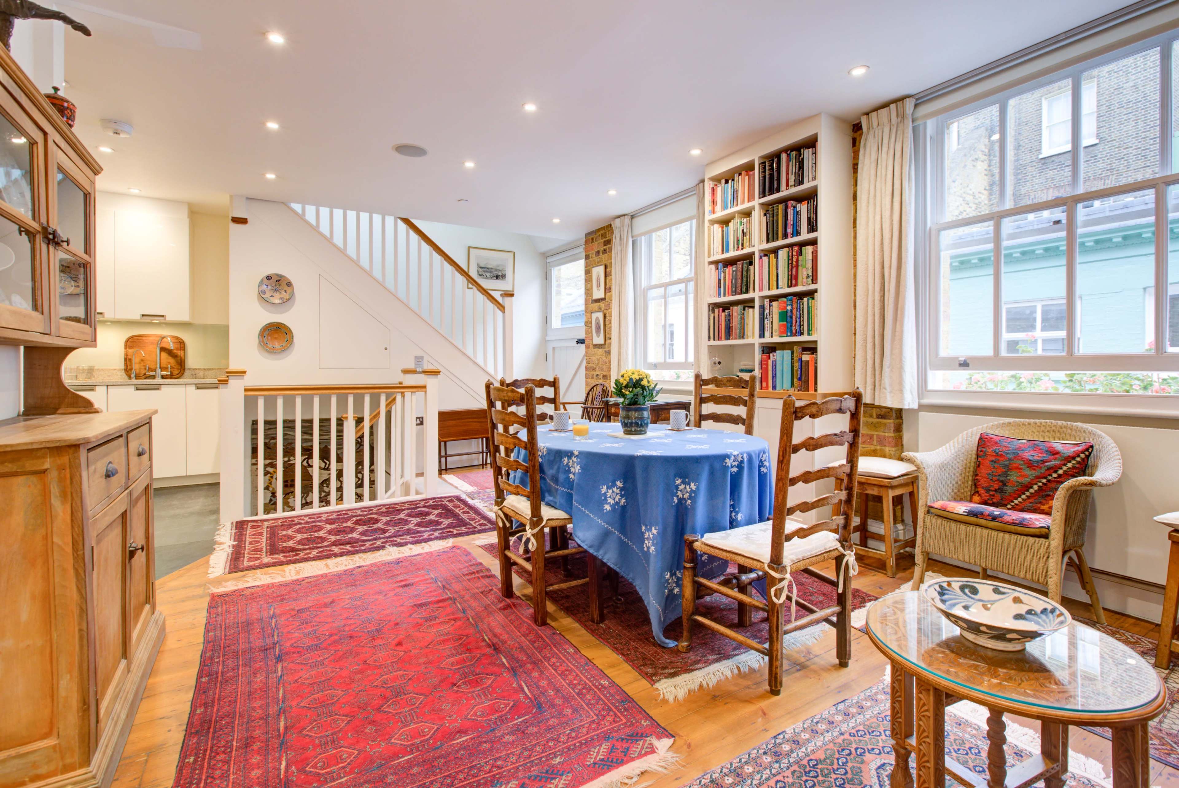 A dining area with a table set for six, surrounded by wooden chairs and adorned with patterned rugs, alongside a bookshelf and a staircase.