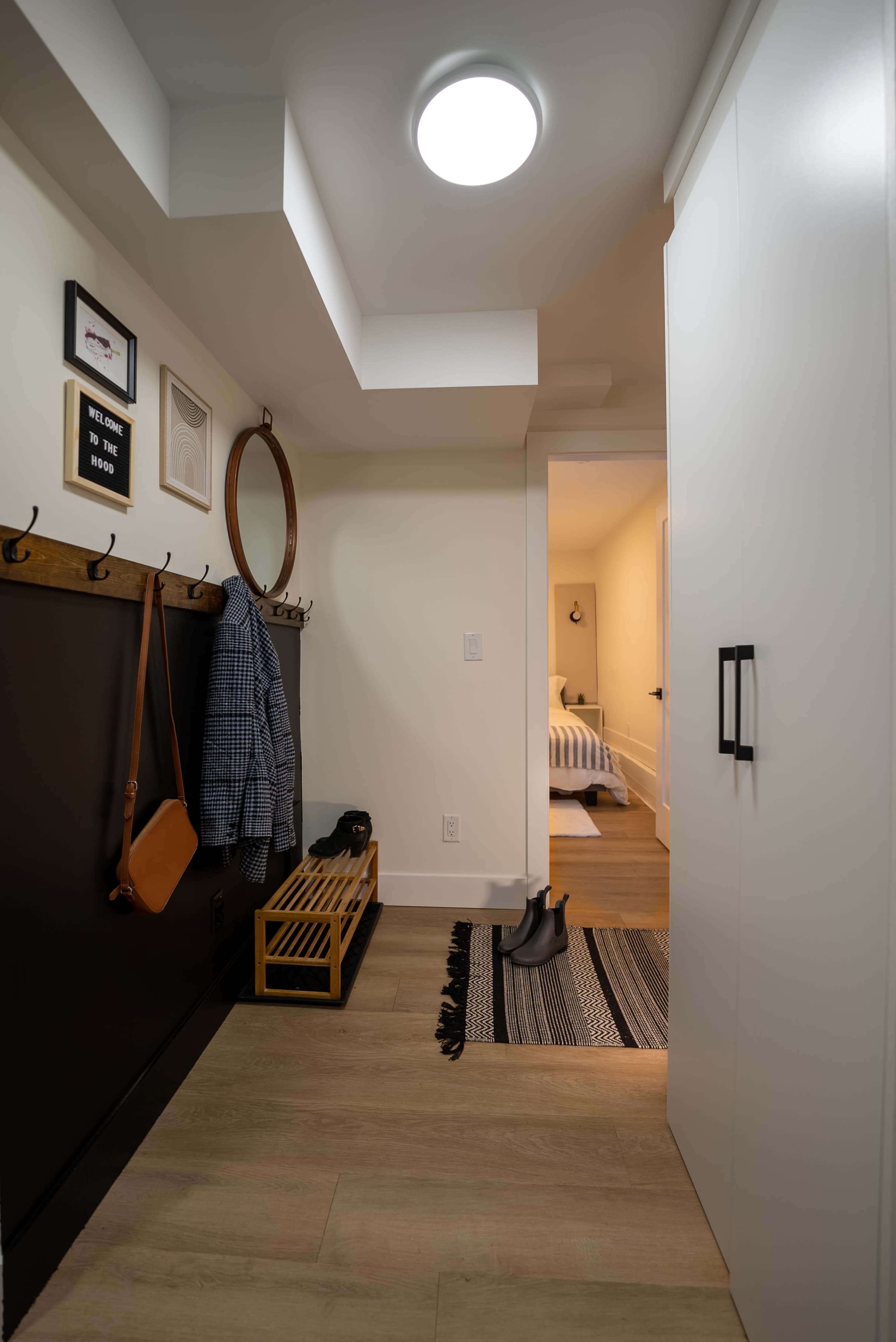 A hallway with a coat rack, a mirror, a wooden bench, and a rug, leading to a room with a bed in the background.