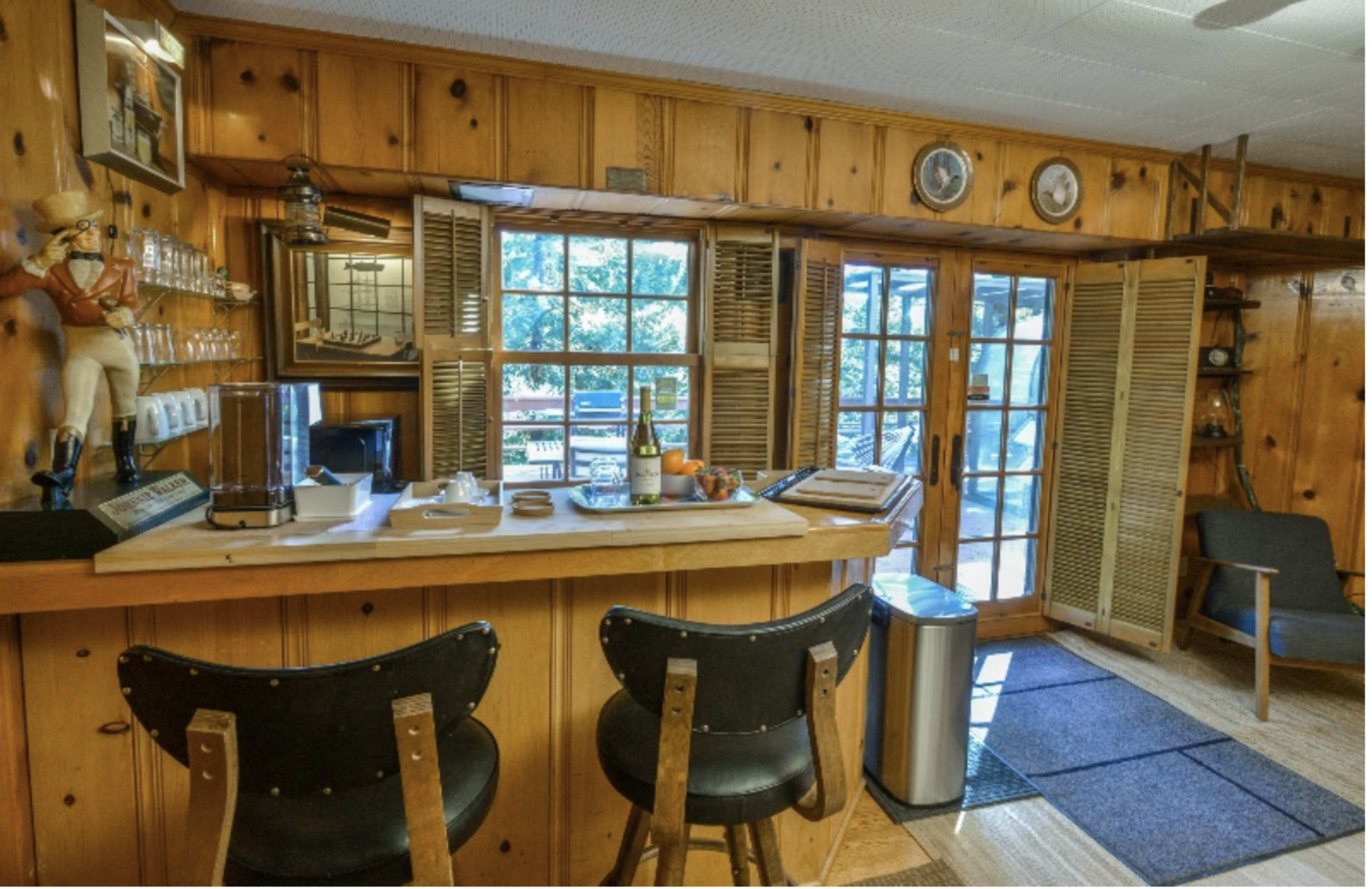 A wooden-paneled room with a bar counter, two black stools, a large set of windows, and a door leading to an outdoor area.