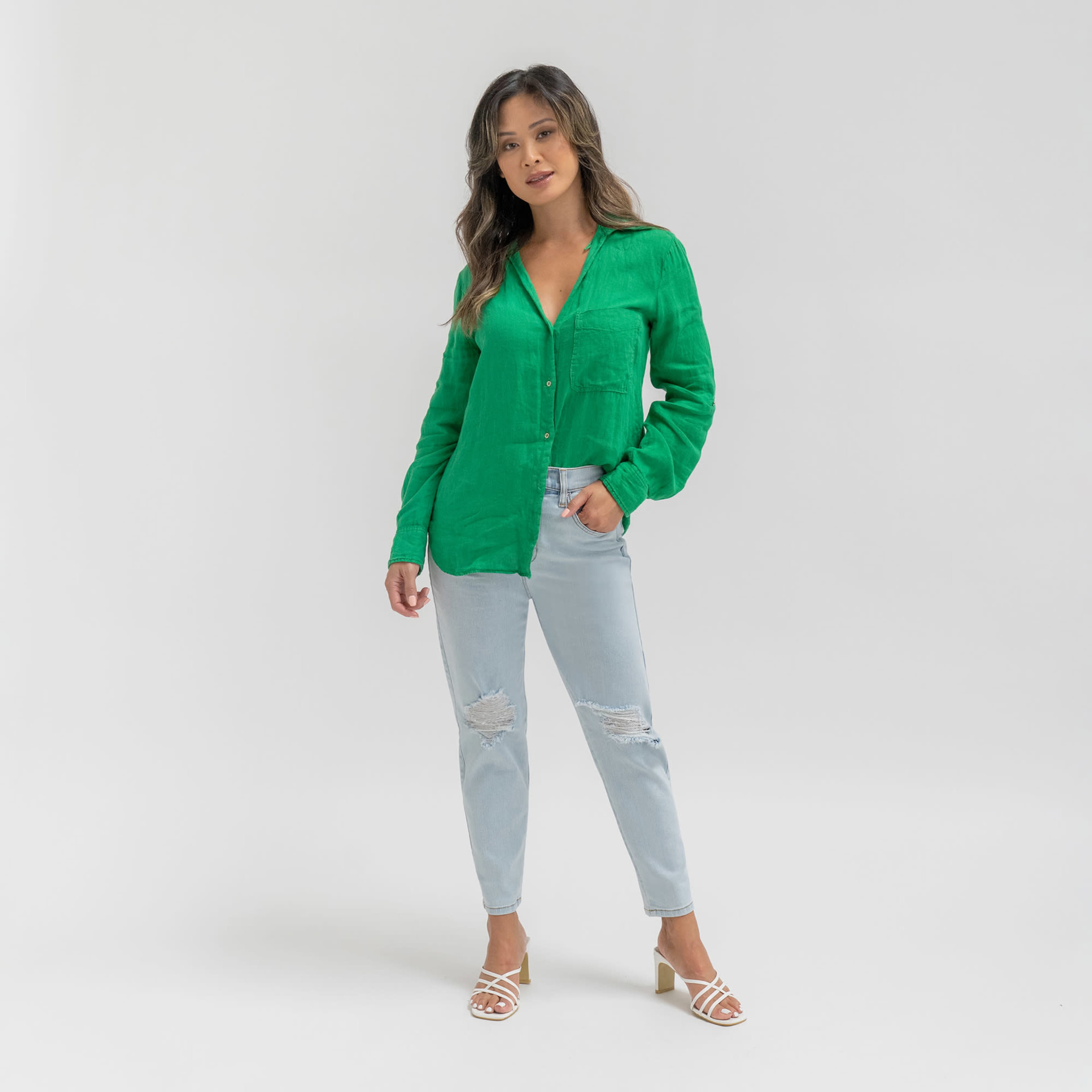 A woman stands against a plain white background, wearing a green button-up shirt and light blue ripped jeans.