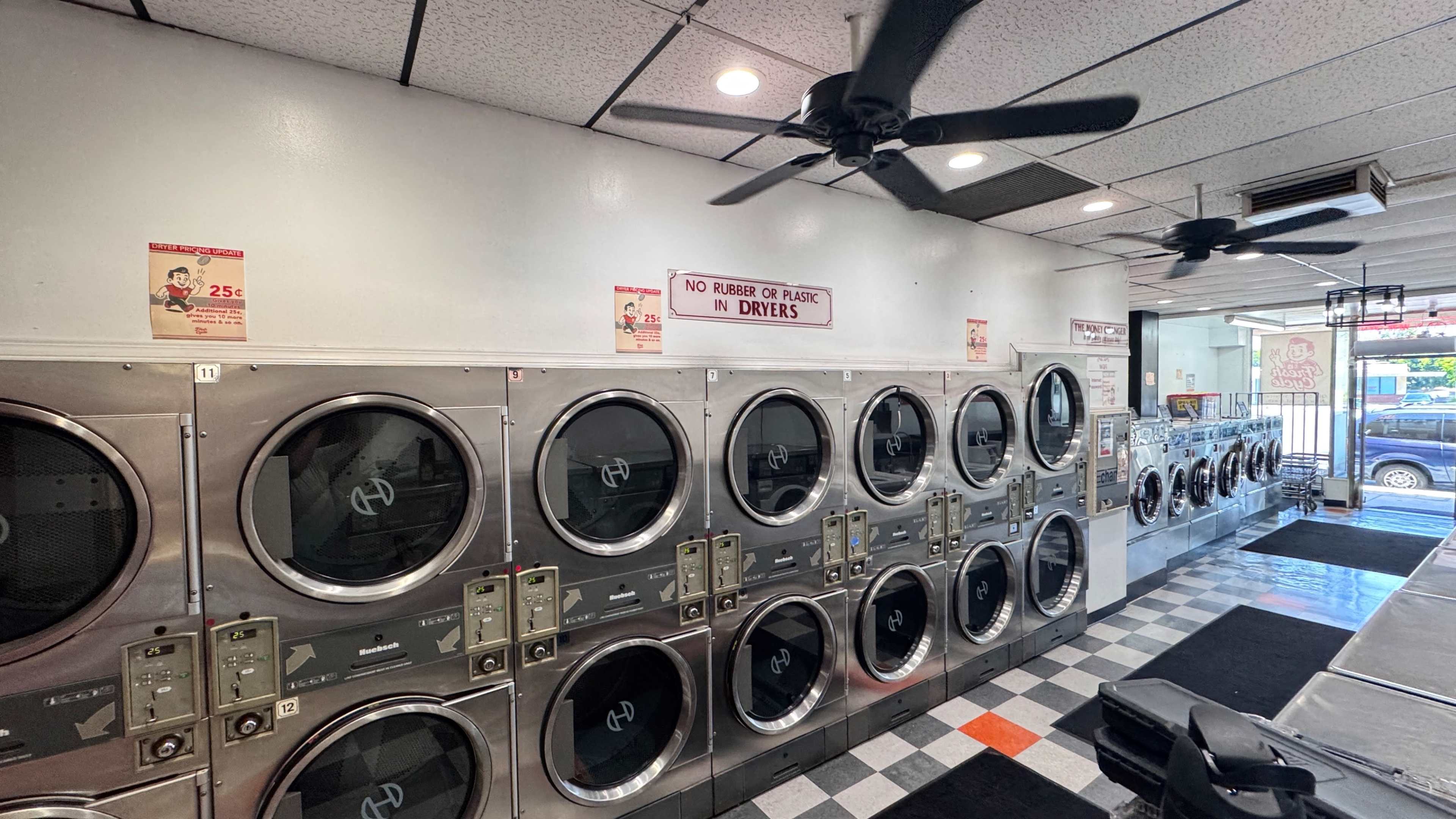 The image shows a laundromat with rows of washing machines and dryers, featuring a clean layout and ceiling fans.