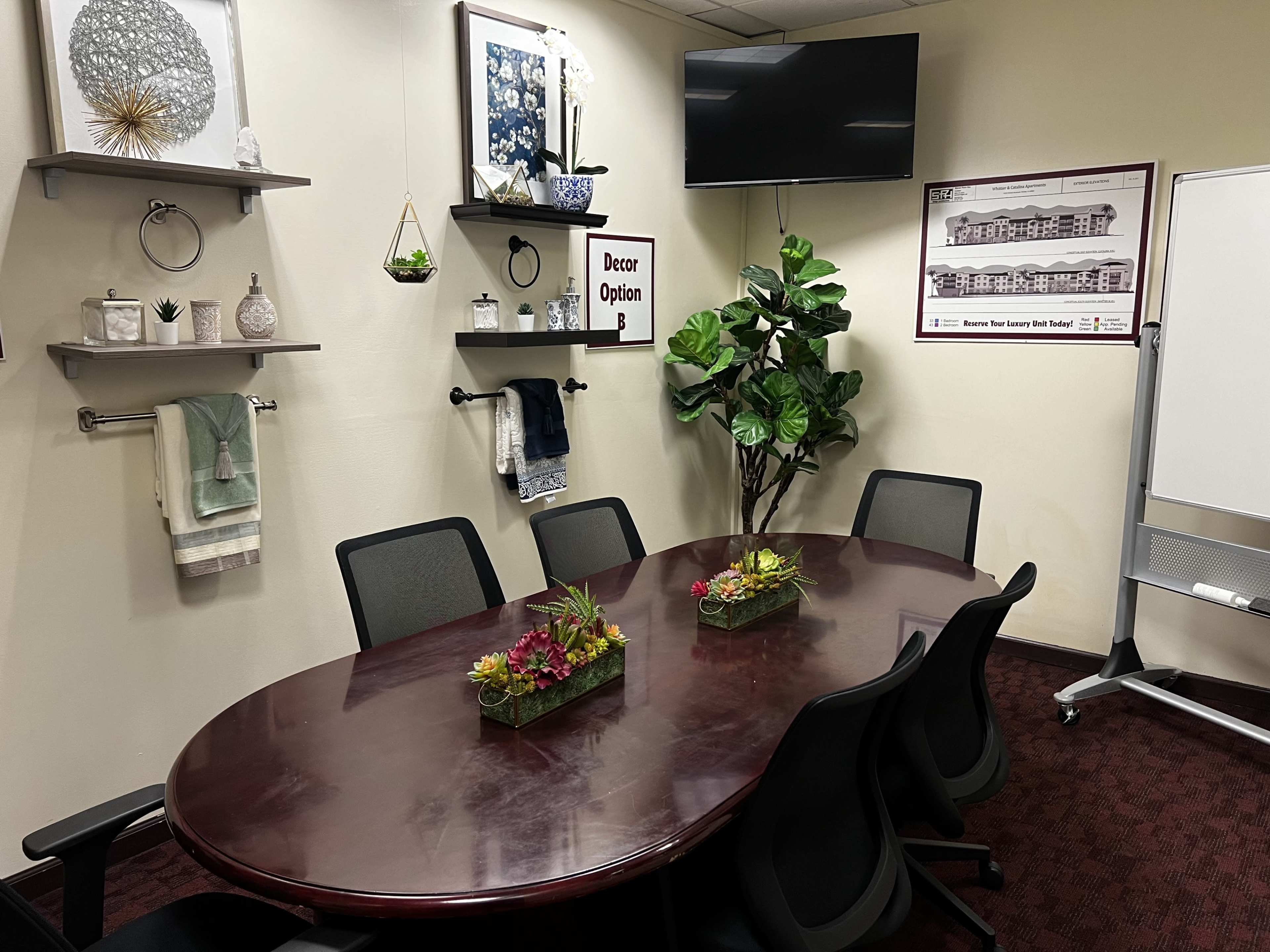 A conference room with a brown oval table surrounded by five black chairs, decorated with potted plants and art on the walls.