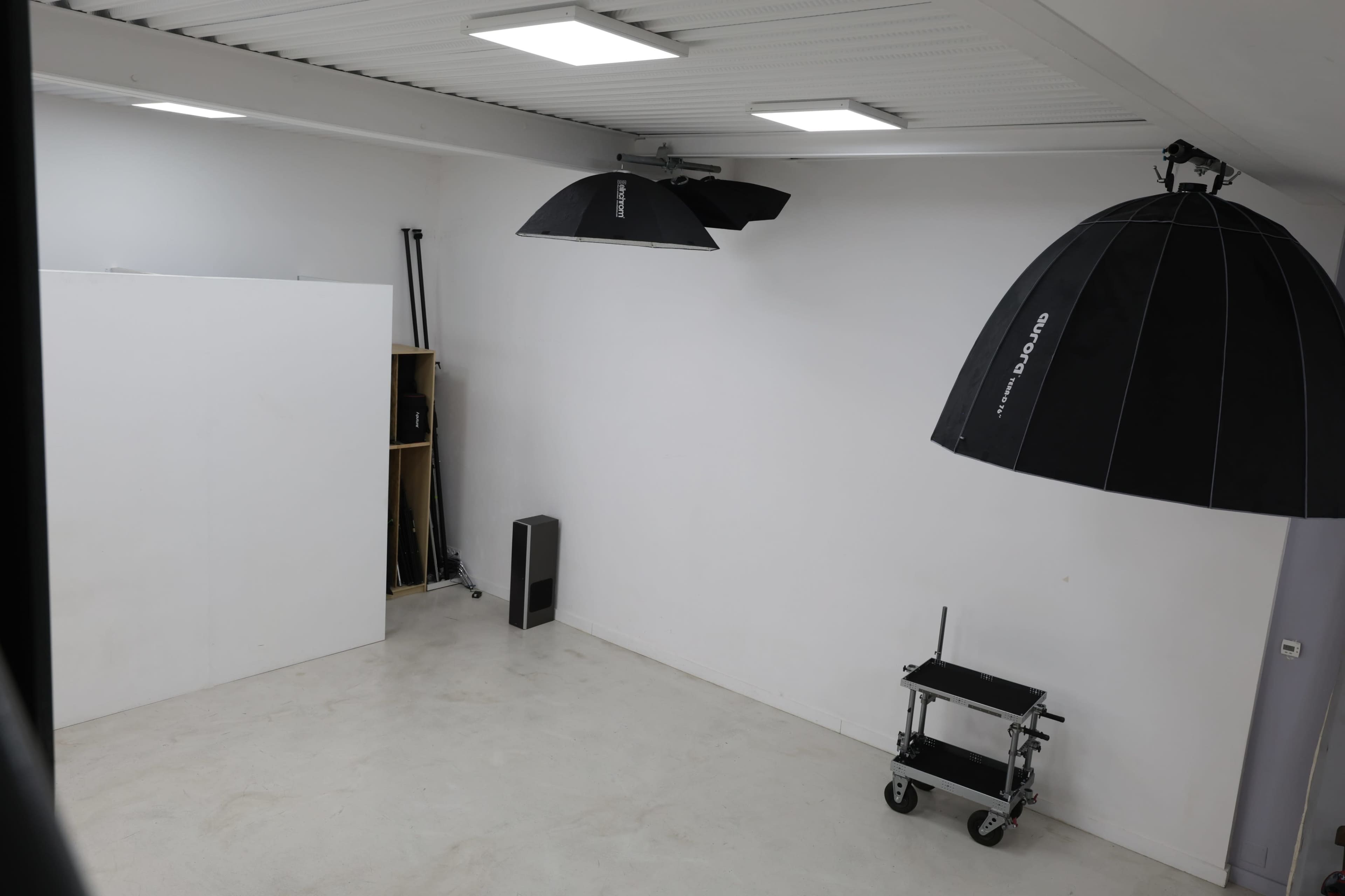 The image shows a minimalist studio space with a white wall, ceiling lights, two light umbrellas hanging, and a small cart in a corner.