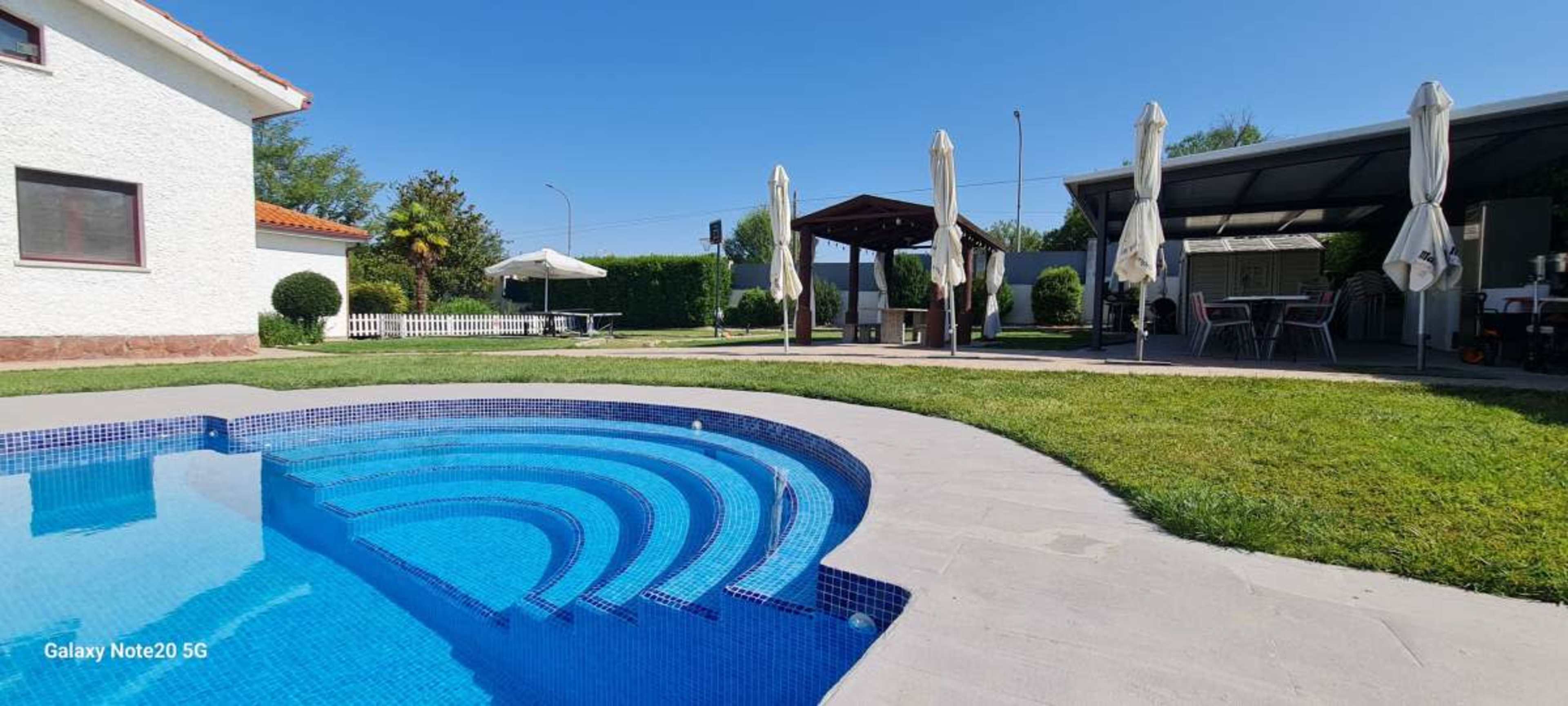 The image shows a backyard with a swimming pool featuring curved steps, a gazebo, outdoor seating, and well-maintained grass under a clear blue sky.