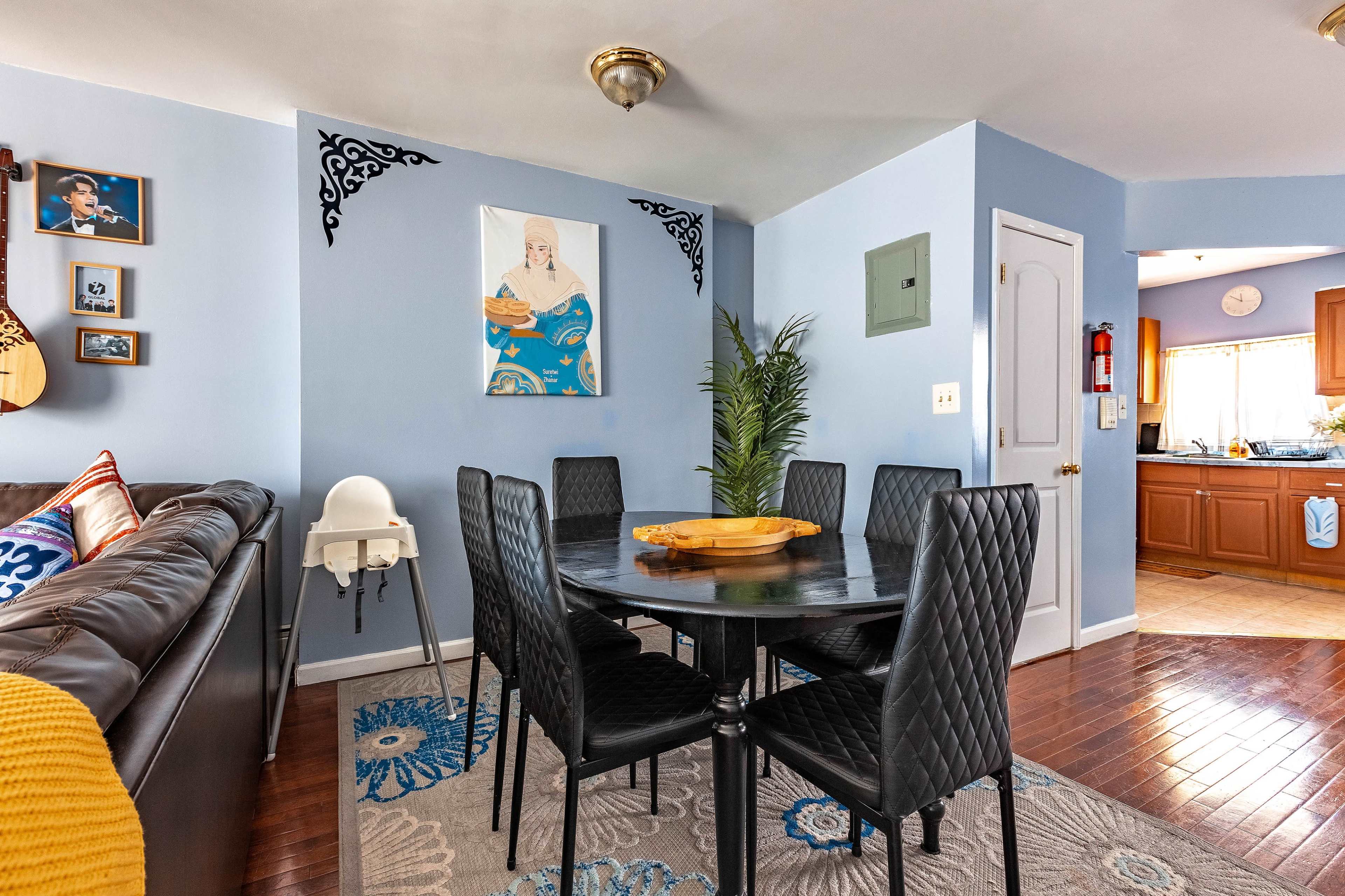 The image shows a dining area with a round black table surrounded by six black chairs, a blue wall with family photos, and a kitchen visible in the background.