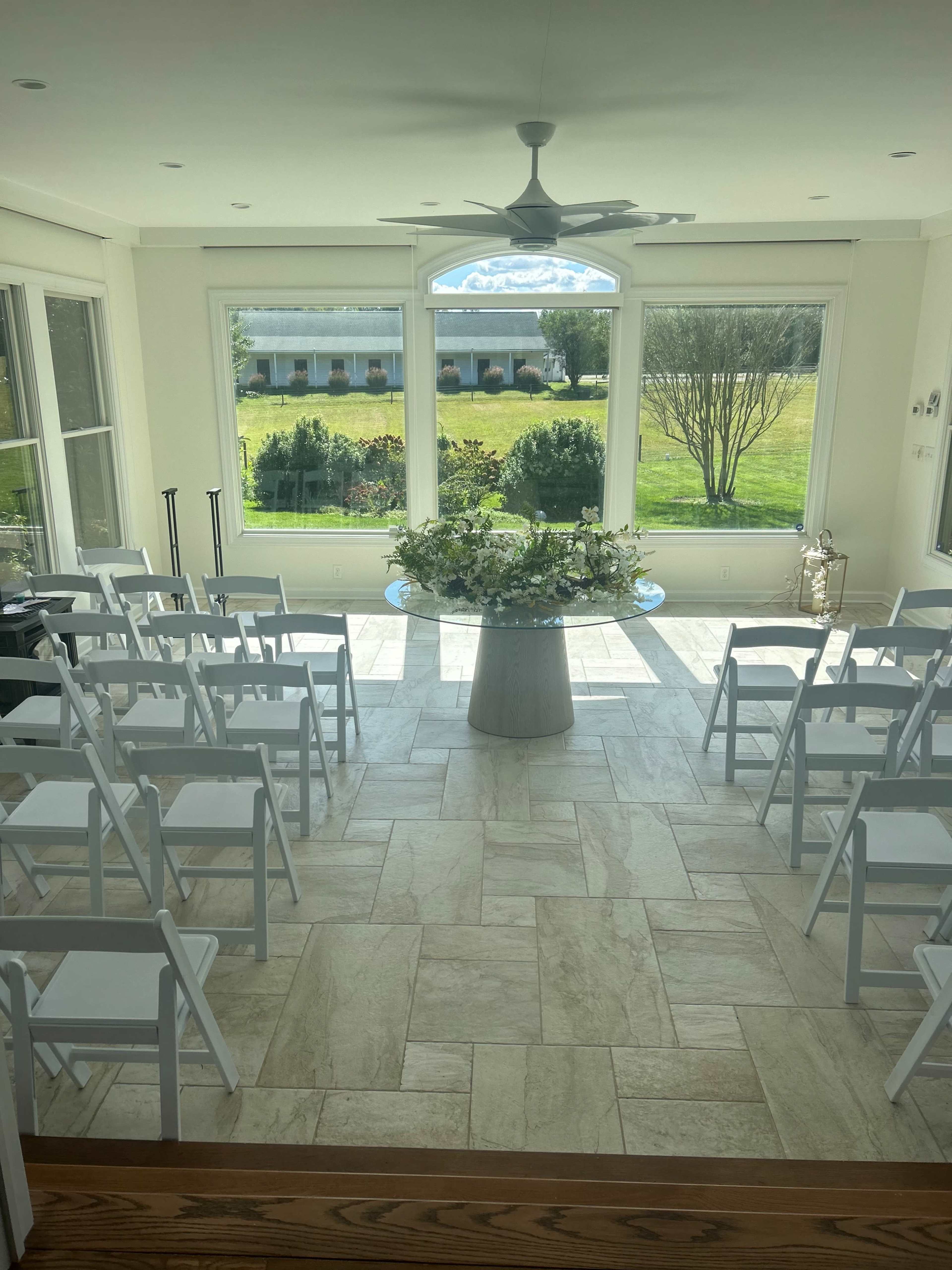 The image shows a bright indoor setting with white folding chairs arranged around a circular floral arrangement in a room with large windows overlooking a green landscape.