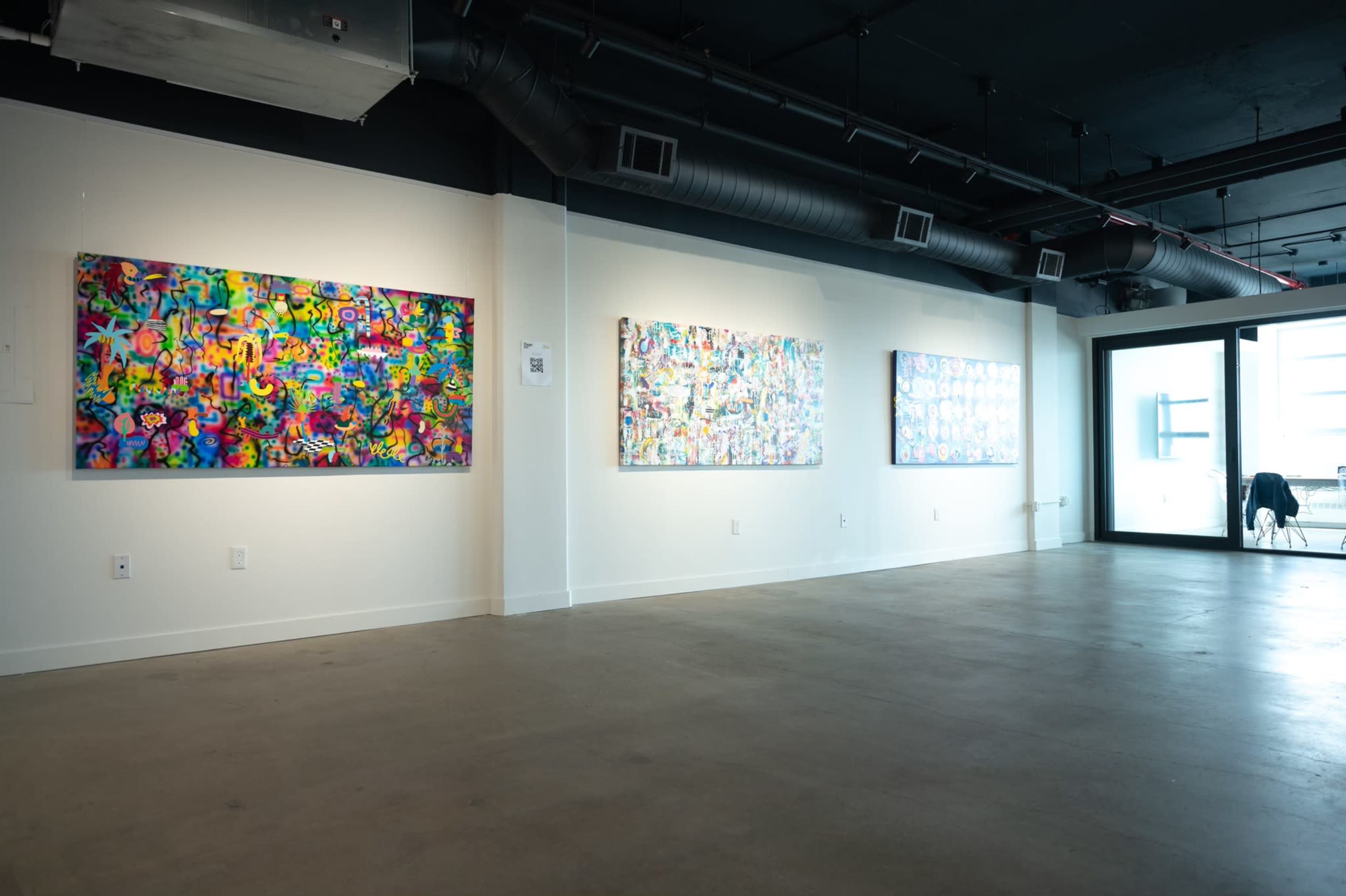 The image shows a gallery space with four colorful abstract paintings displayed on white walls.