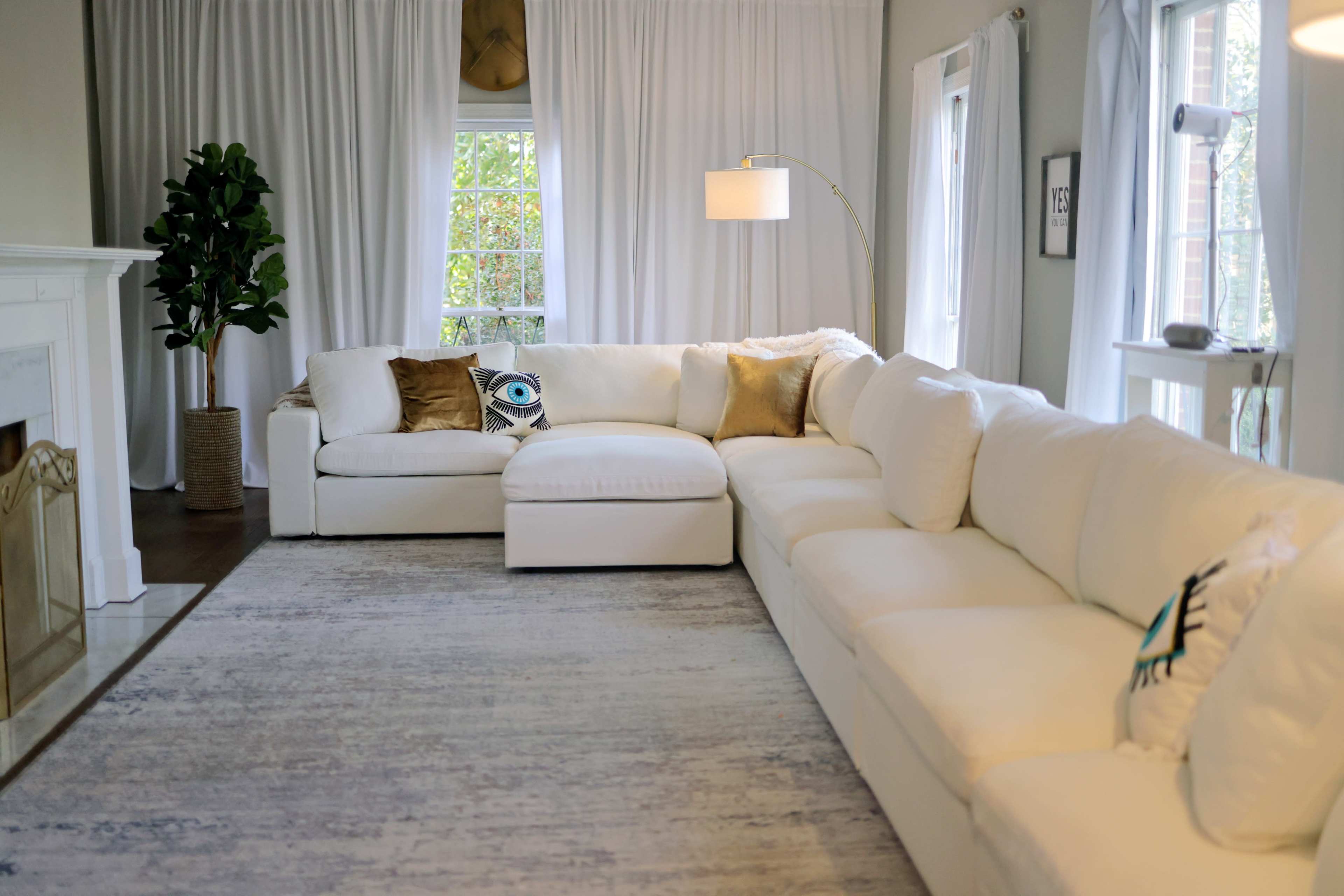 The image shows a light-colored, cozy living room featuring a large sectional sofa, a floor lamp, and a decorative plant near the window.