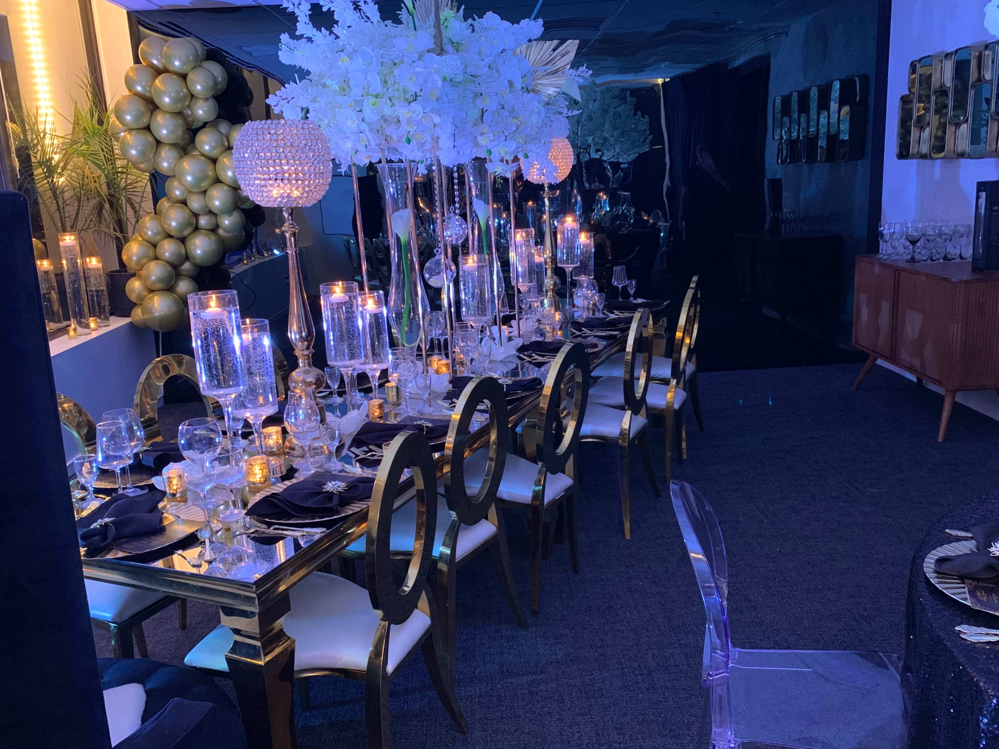 A formal dining setup features a long table adorned with elegant glassware, ornate candle holders, and floral arrangements, surrounded by gold and transparent chairs in a dimly lit room.