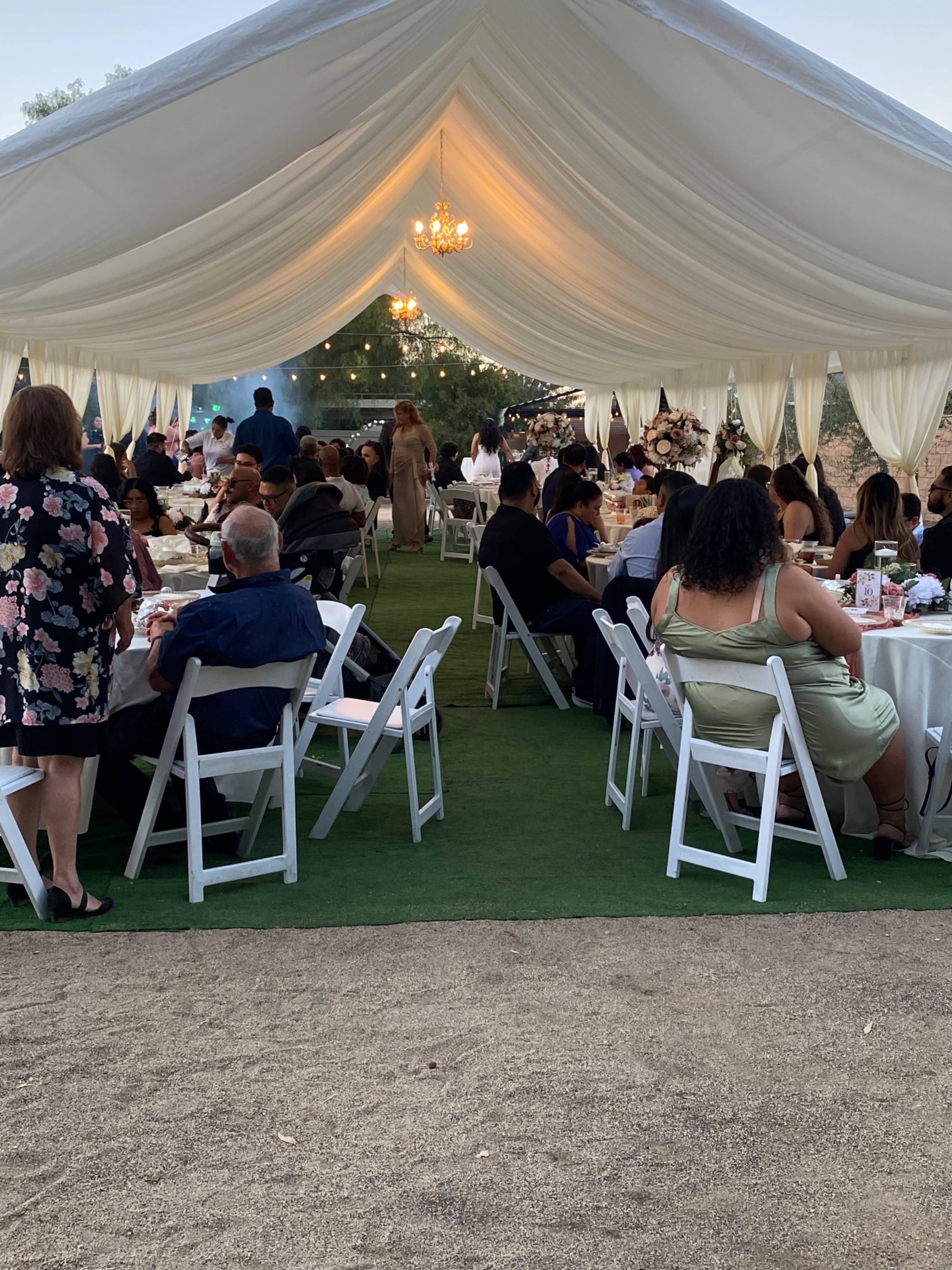 A large outdoor tent is set up for a gathering, with guests seated at round tables adorned with white tablecloths, and decorative elements under a soft, draped ceiling.