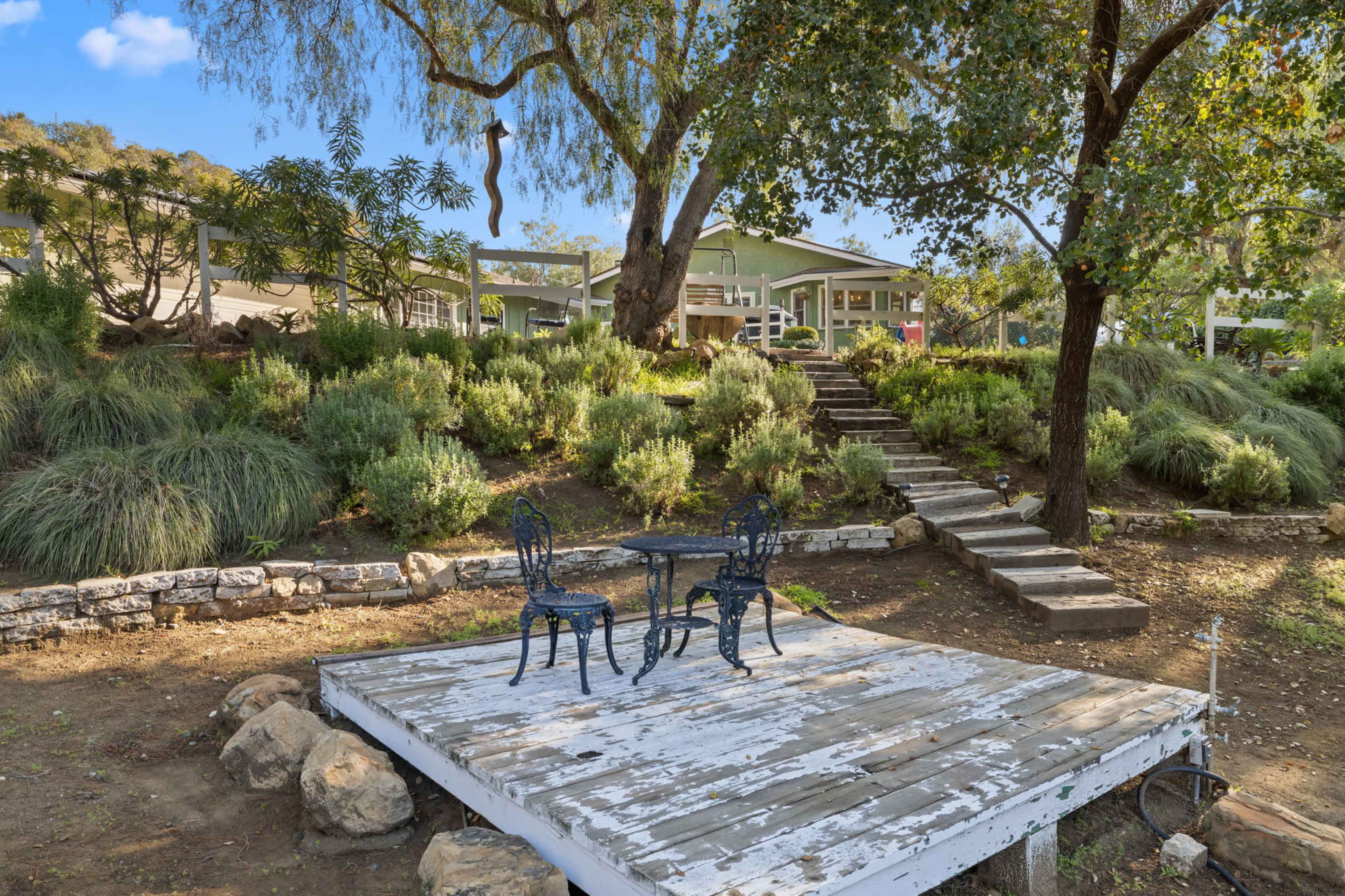 A wooden deck with a black metal table and chairs is situated in a garden area surrounded by trees and shrubs, leading up to a house on a hillside.