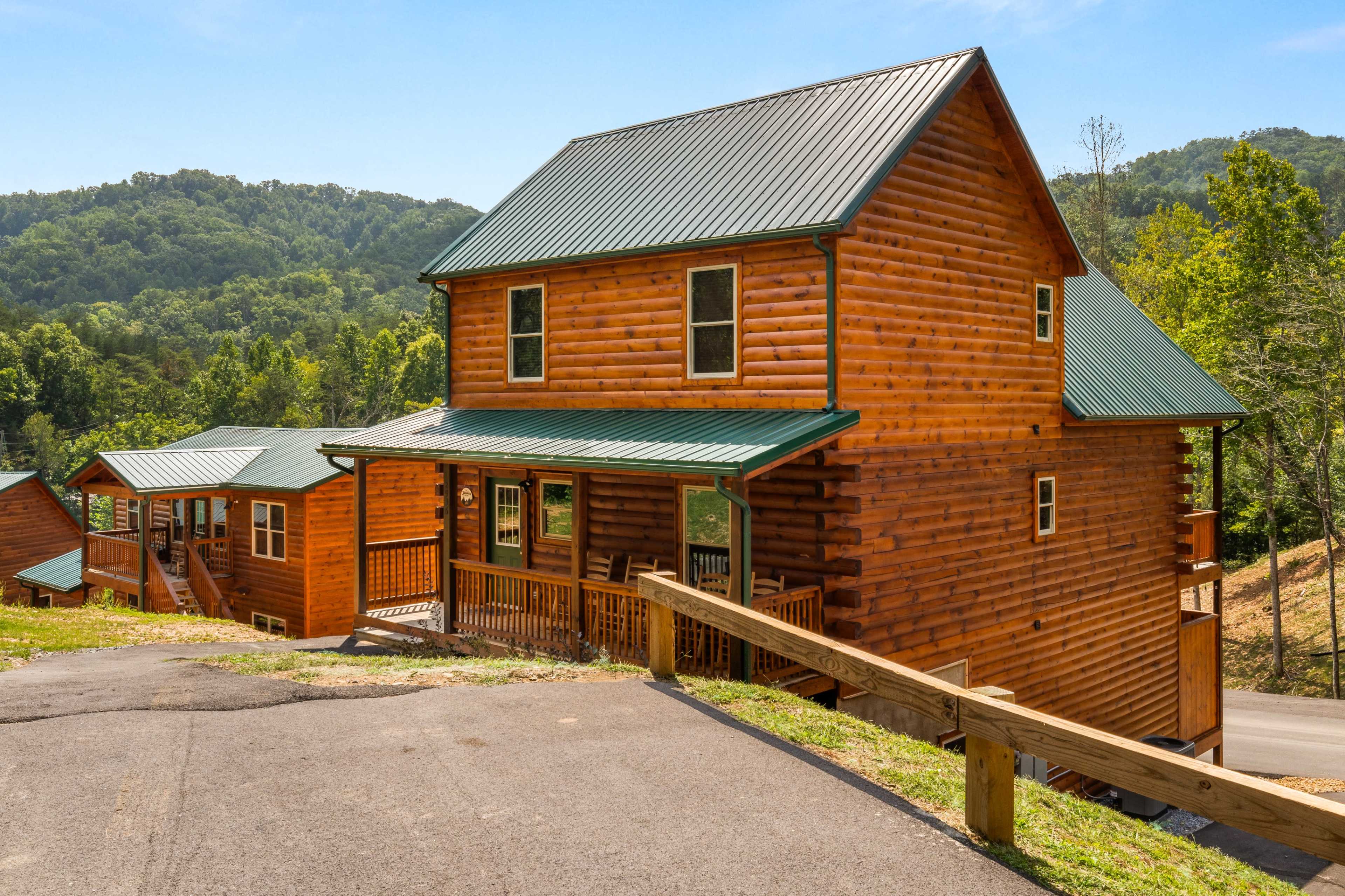 A two-story log cabin with a green metal roof is situated on a hillside surrounded by trees and mountains.