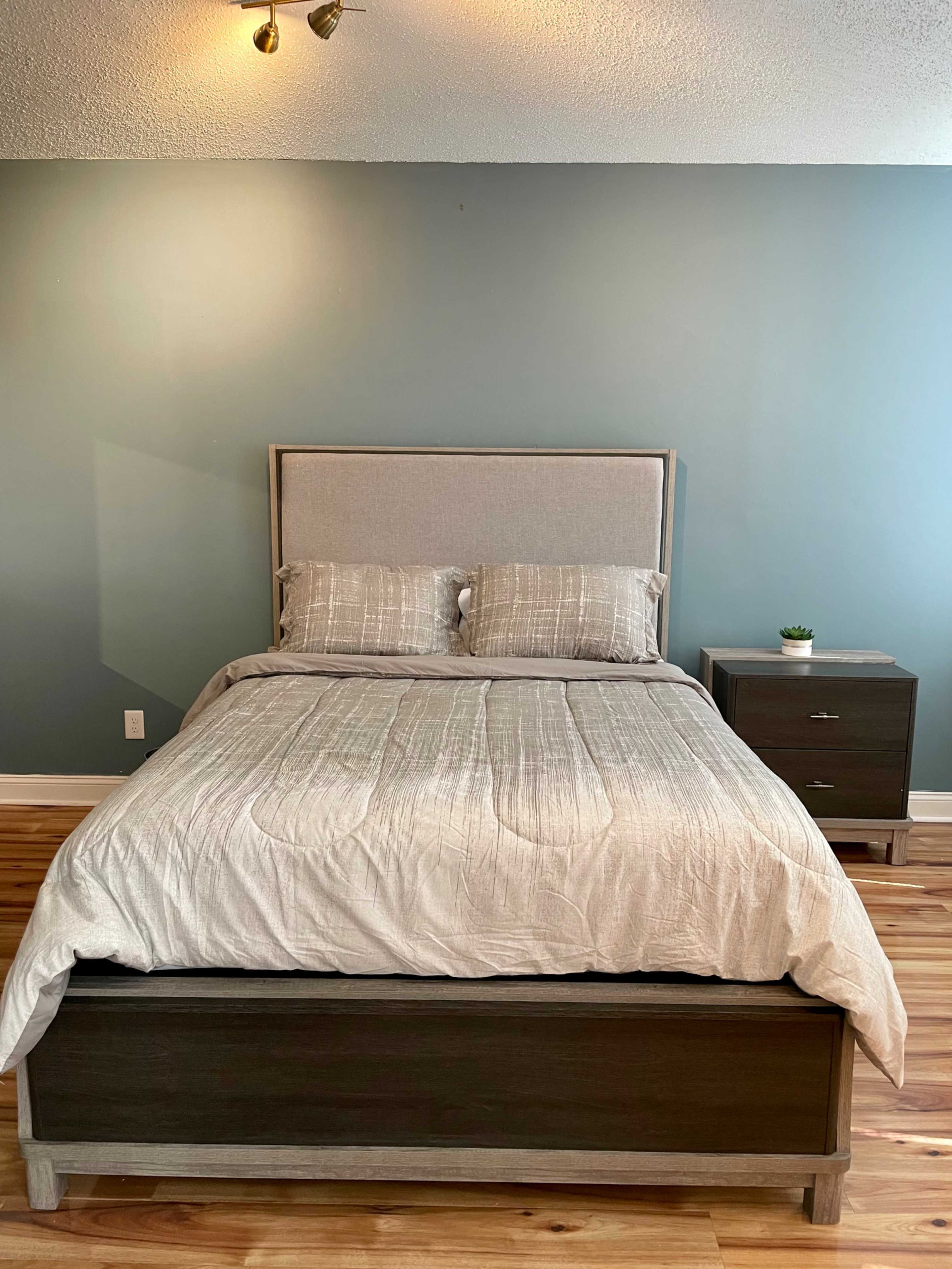 A neatly made bed with a light gray comforter and two pillows, set against a blue wall, accompanied by a nightstand on one side.
