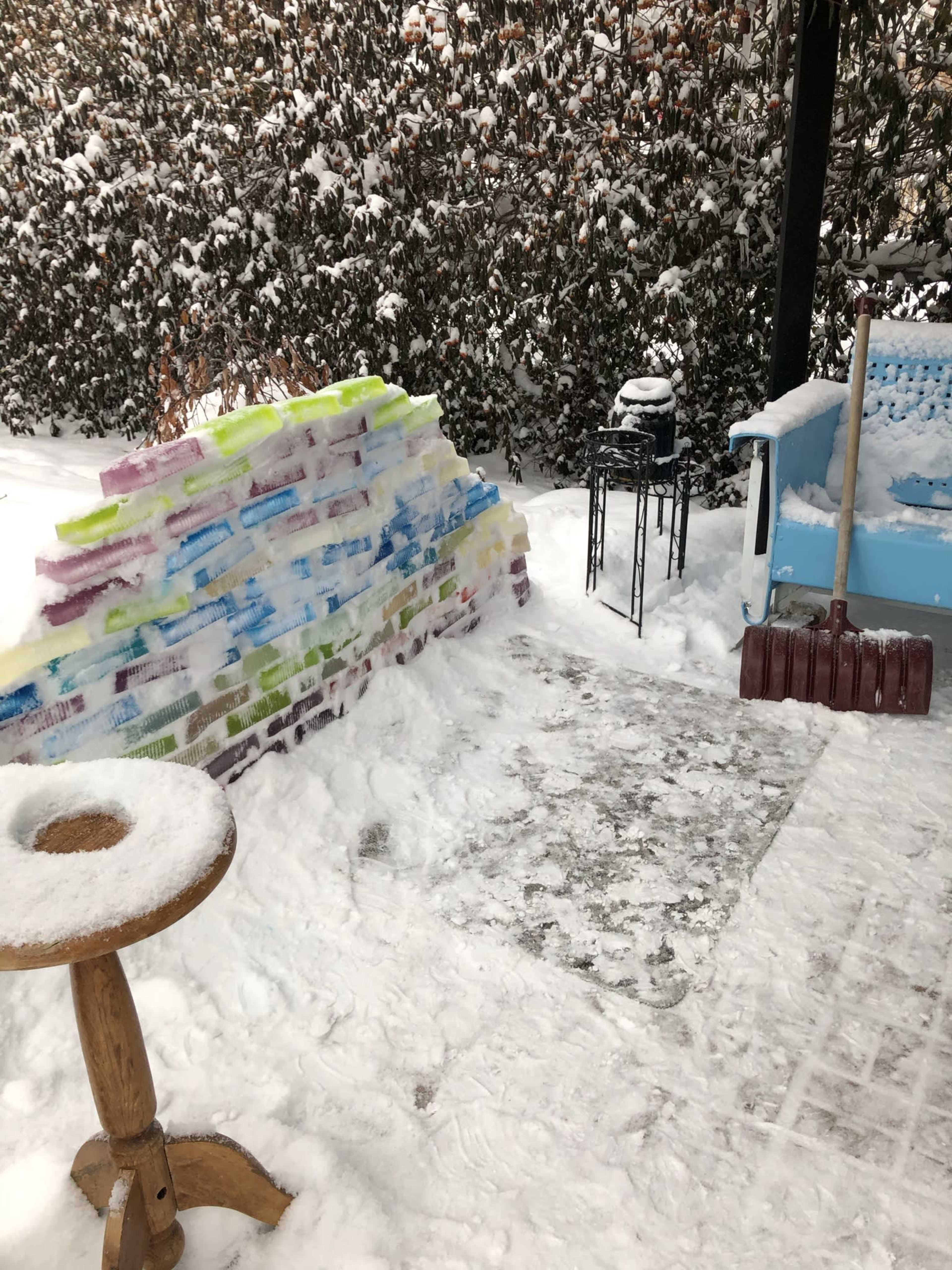 A snow-covered outdoor area with a colorful, ice brick wall, a small table, and a shovel resting on the ground.