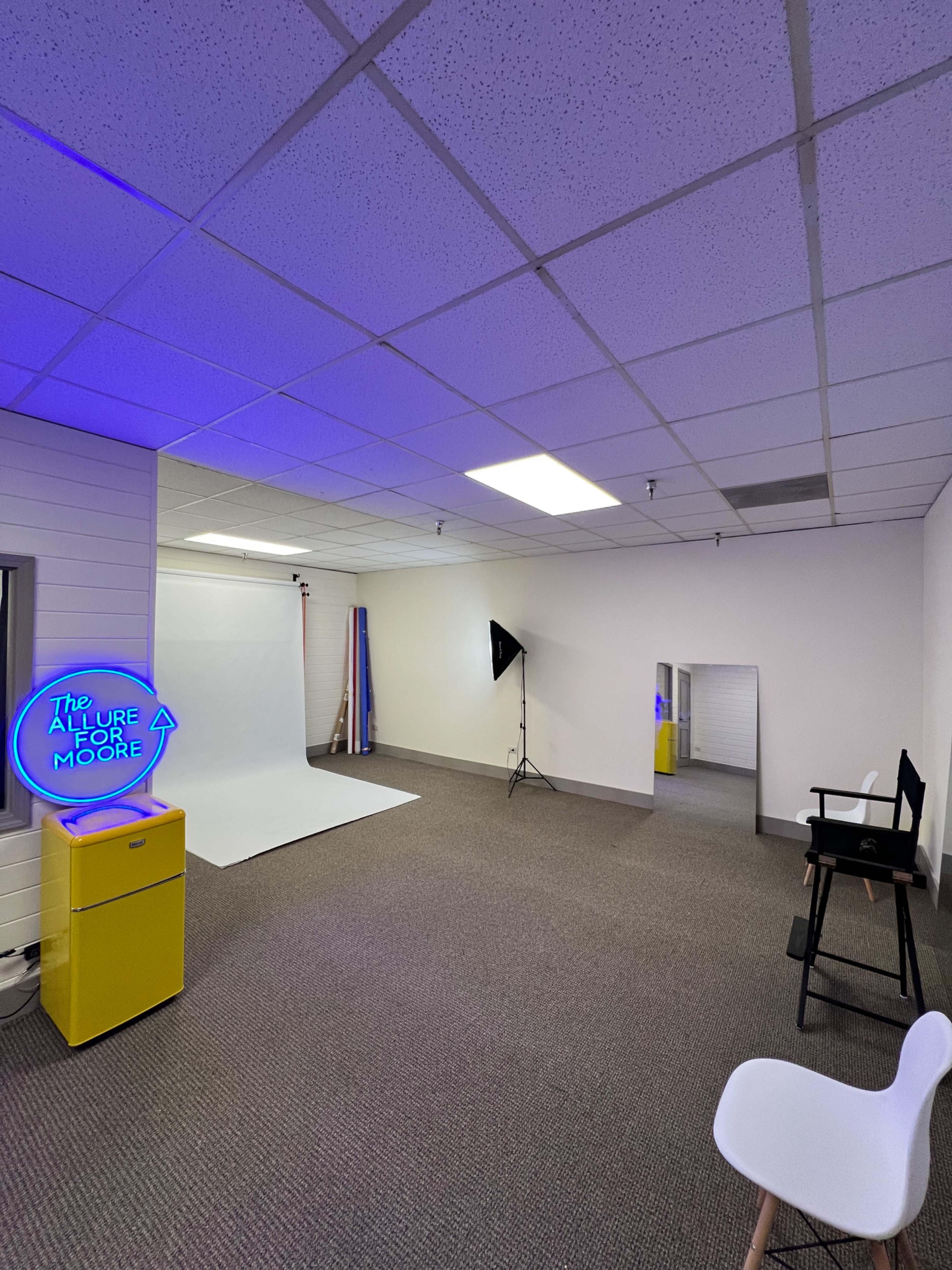 The image shows a spacious room with a white backdrop, a director's chair, a bright yellow cabinet, and purple lighting.
