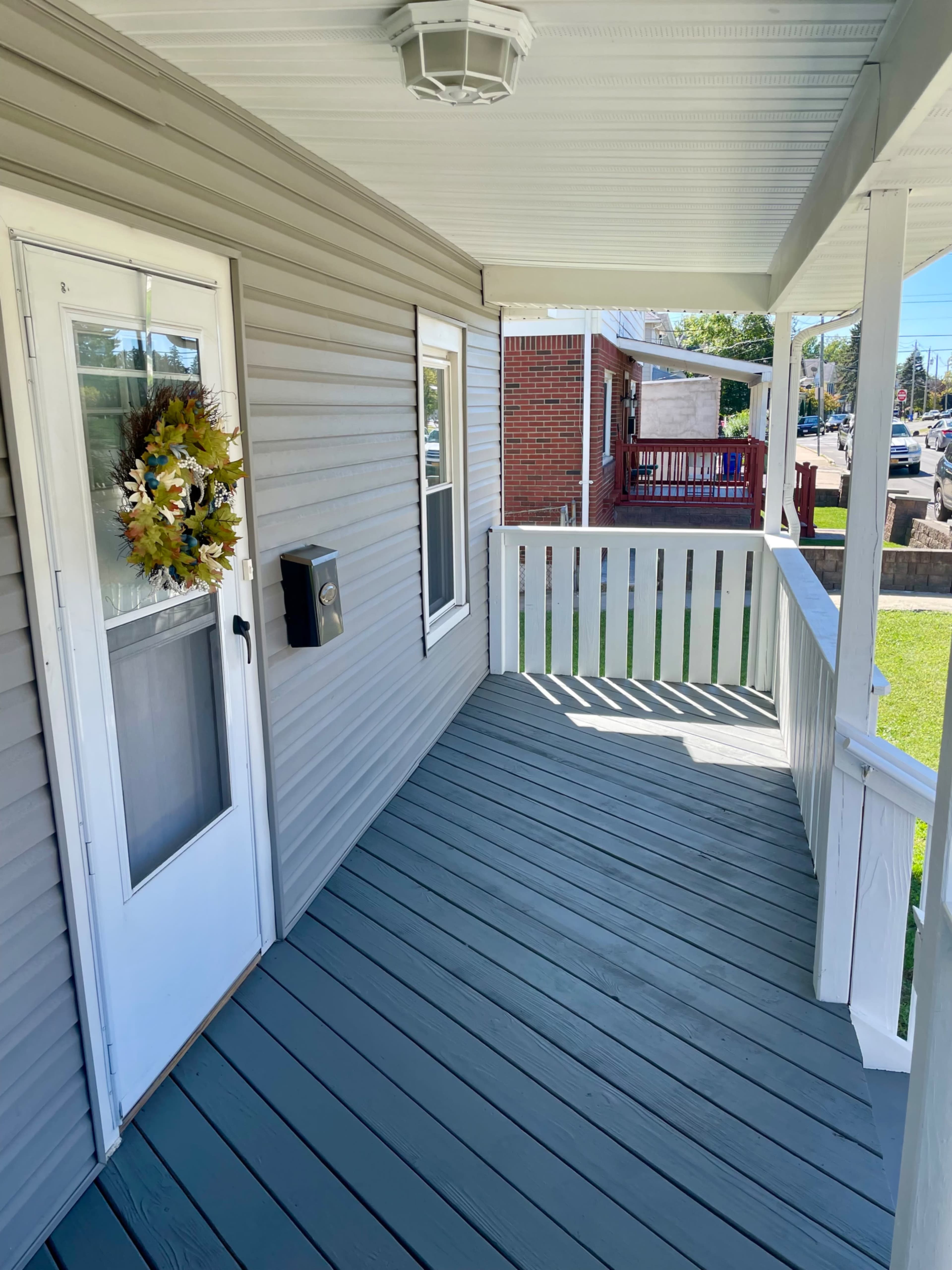 The image shows a front porch with a white door, a decorative wreath, a mailbox on the side, and a gray floor, surrounded by a white railing.