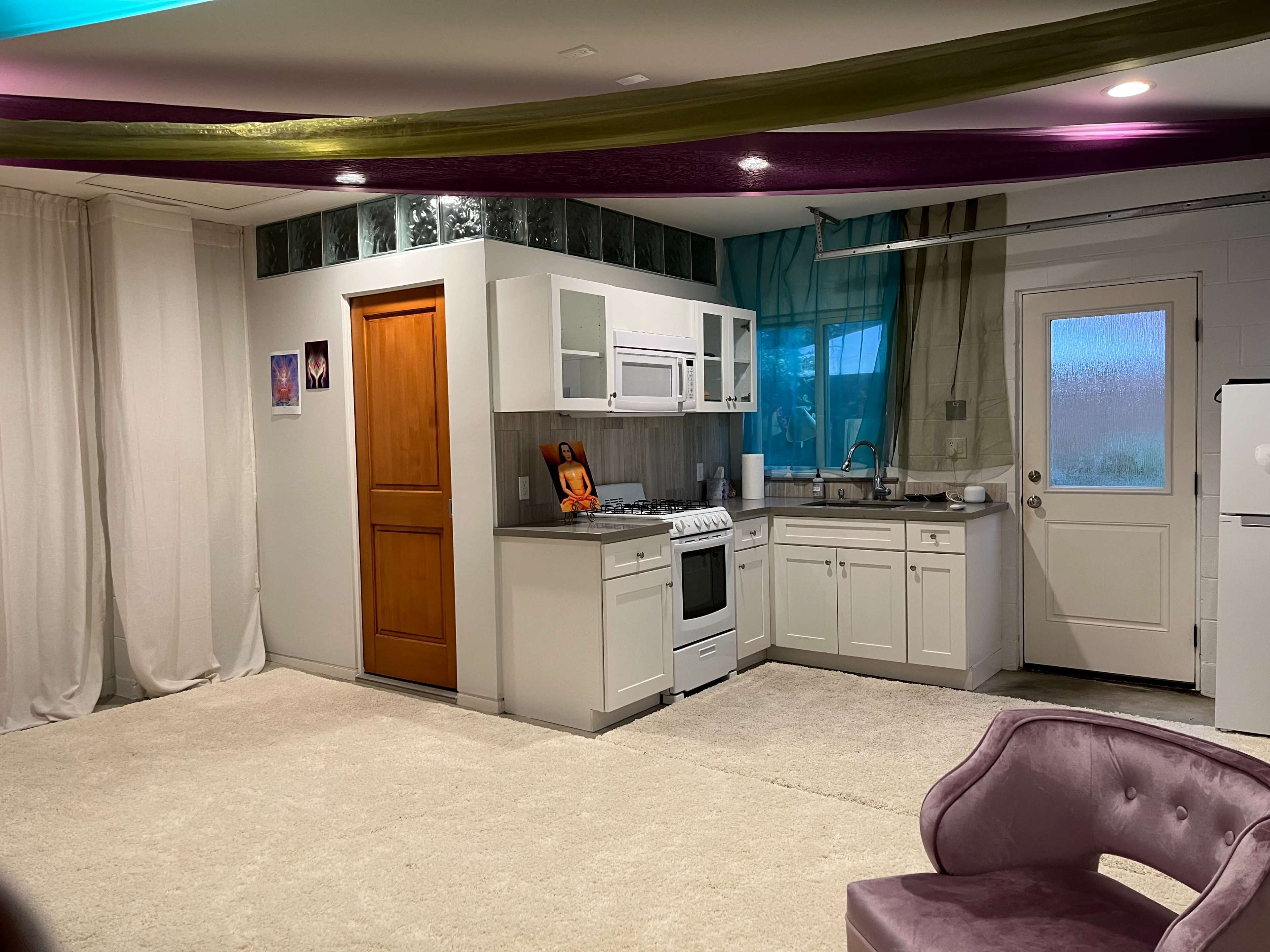The image shows a compact kitchen area with white cabinets, a stove, and a door leading outside, set against a backdrop of draped fabric and a carpeted floor.