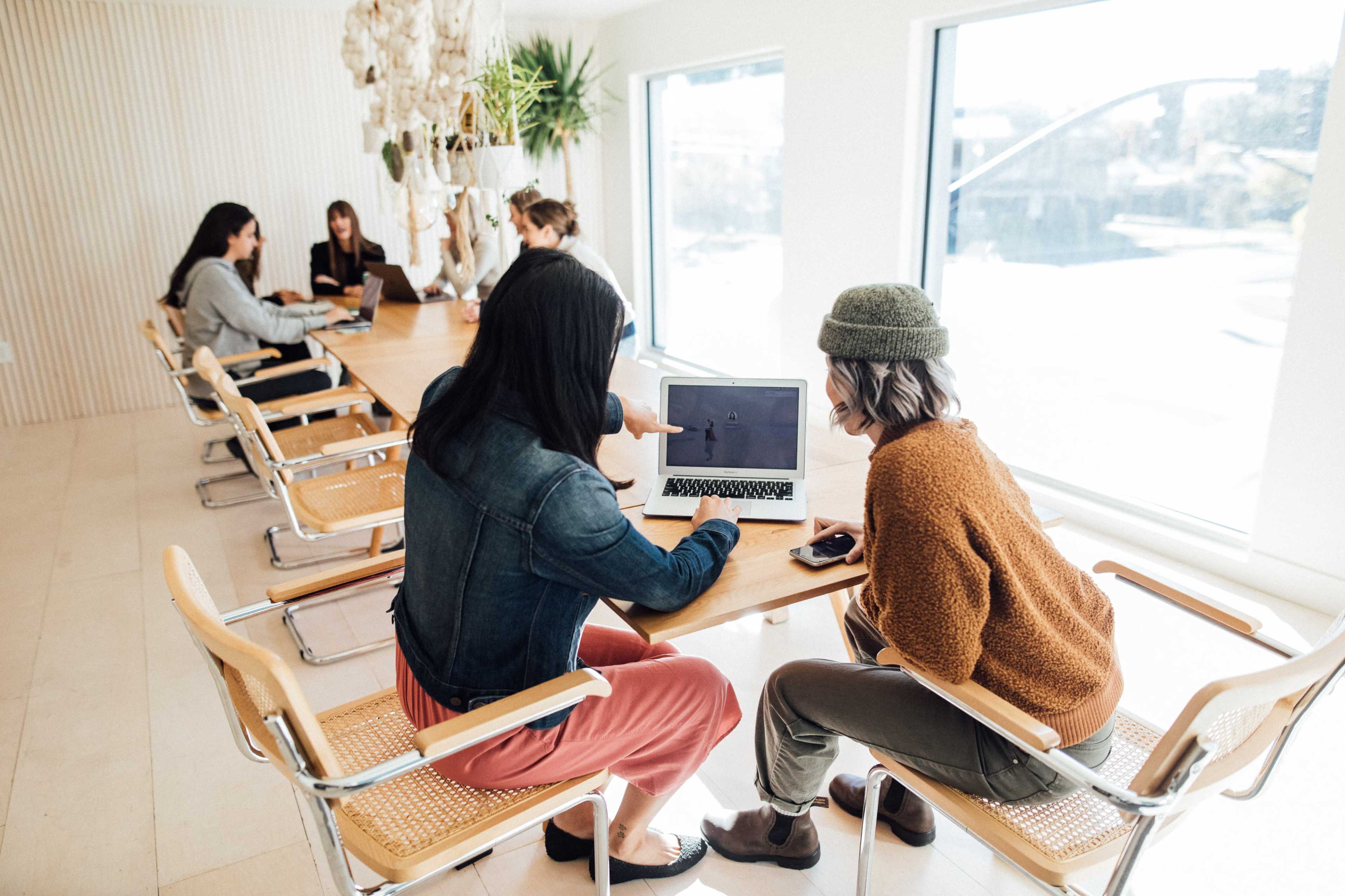 A group of six people works collaboratively at a long wooden table in a modern conference room with large windows.