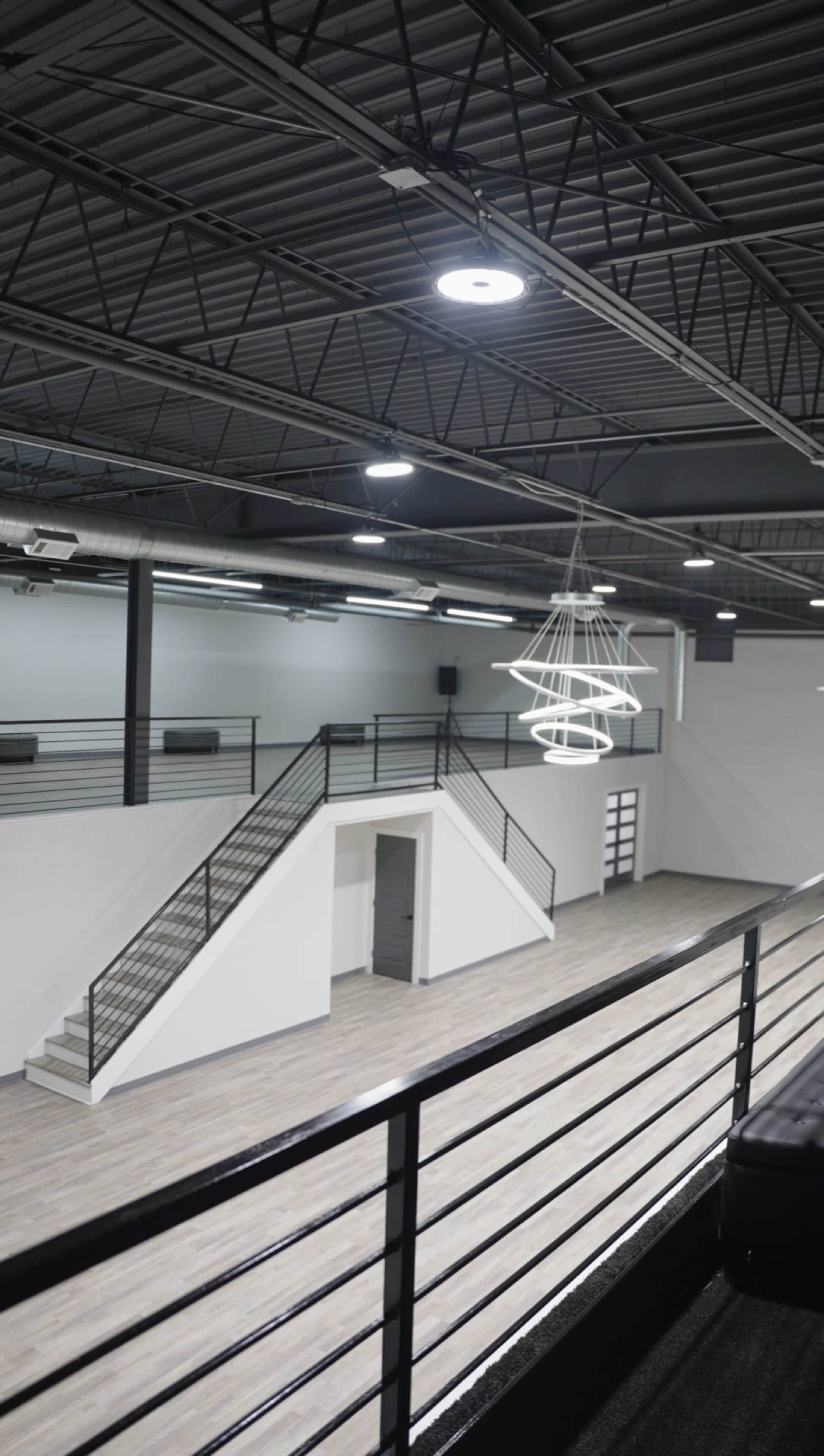 The image shows a modern, spacious interior with a staircase leading to an upper level, featuring sleek railings and a large circular light fixture hanging from the ceiling.