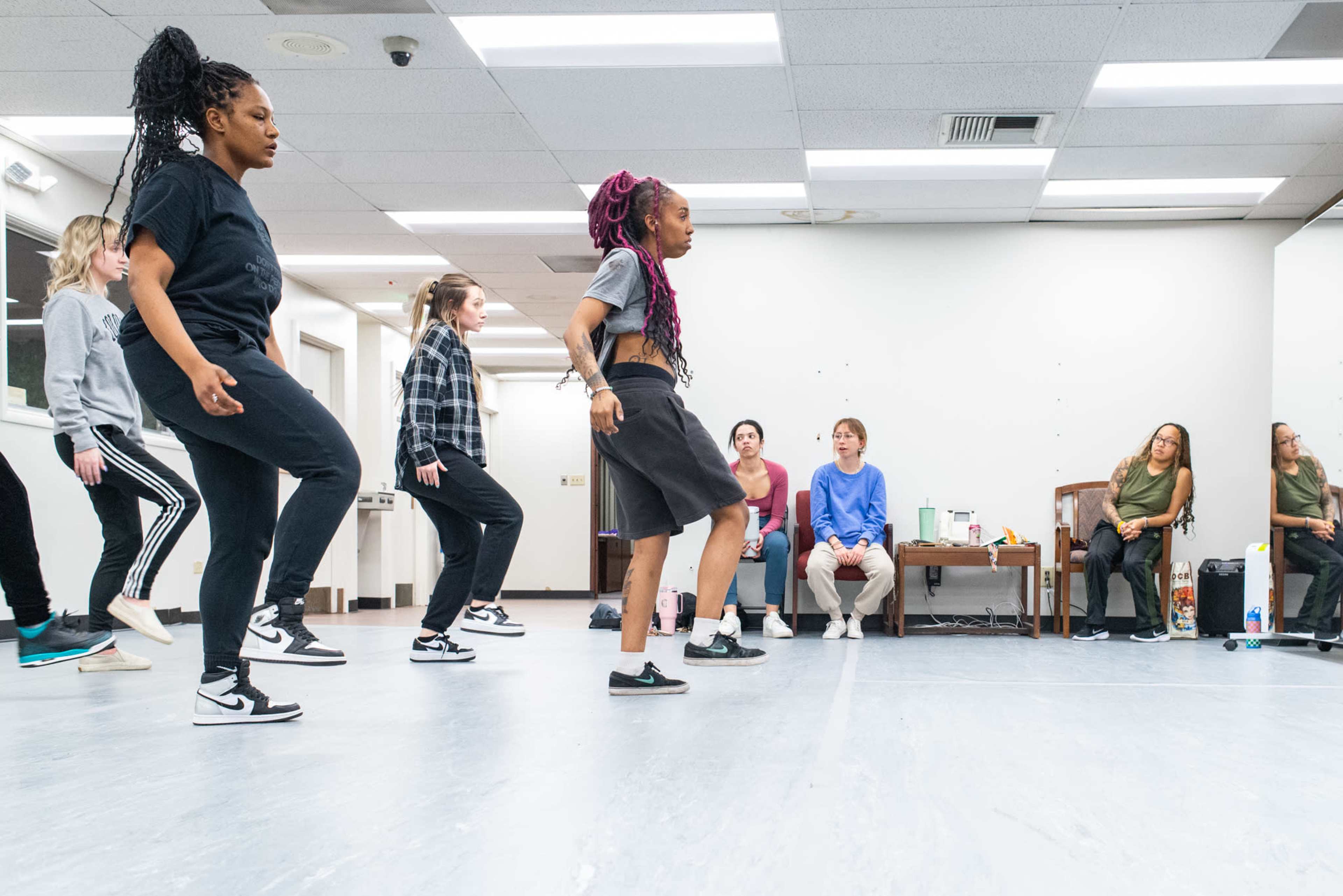 A group of dancers practices footwork in a spacious studio while several observers sit on the side.