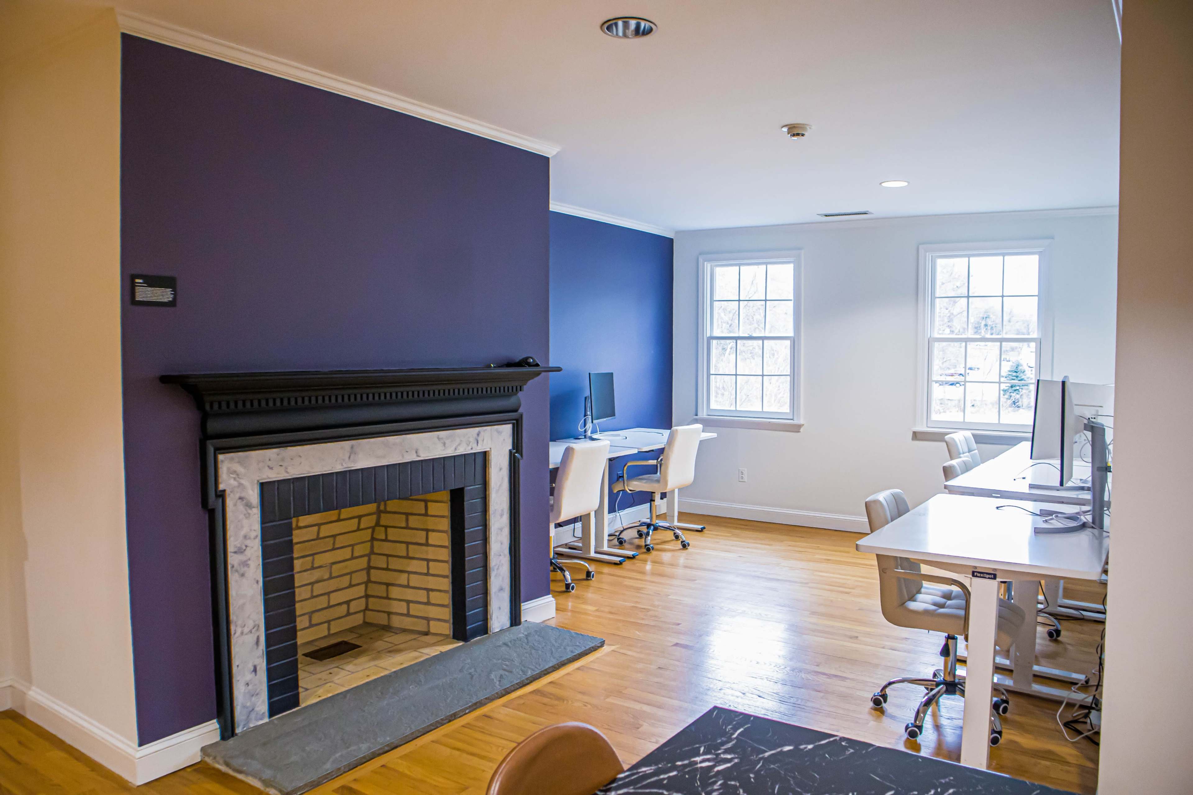 The image shows a modern office space featuring a brick fireplace on a purple wall and several workstations with computers.