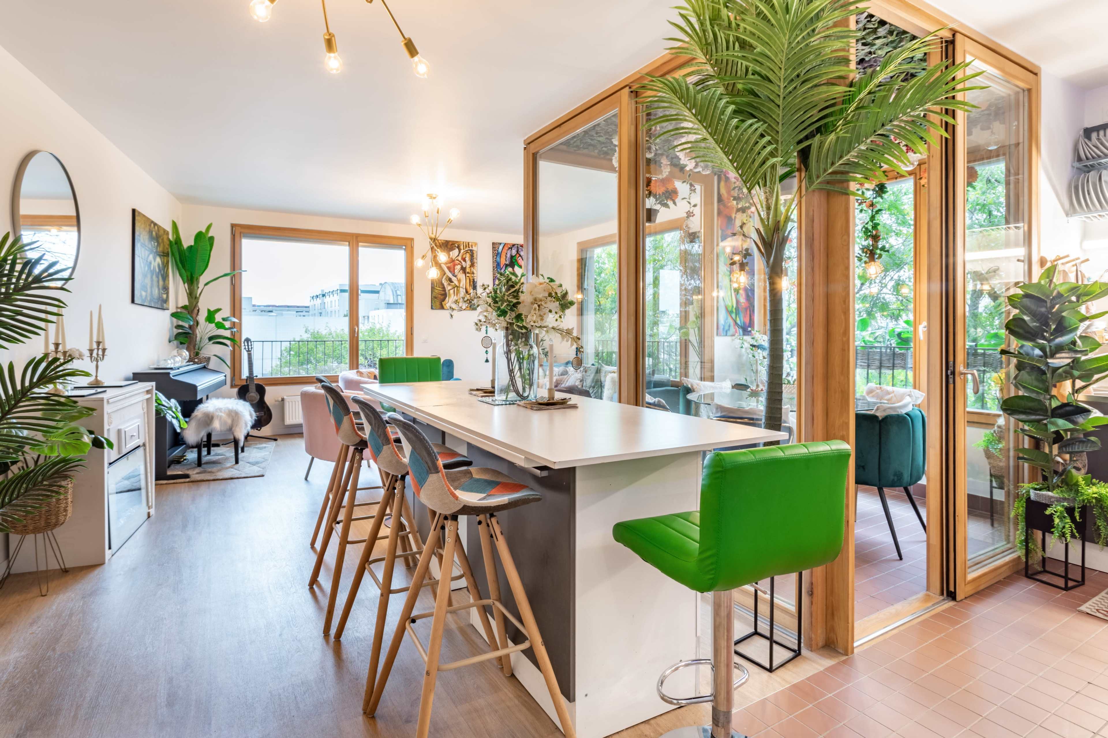 A modern open-concept living space features a kitchen island with bar stools, a dining area, and a glass-enclosed room filled with plants.