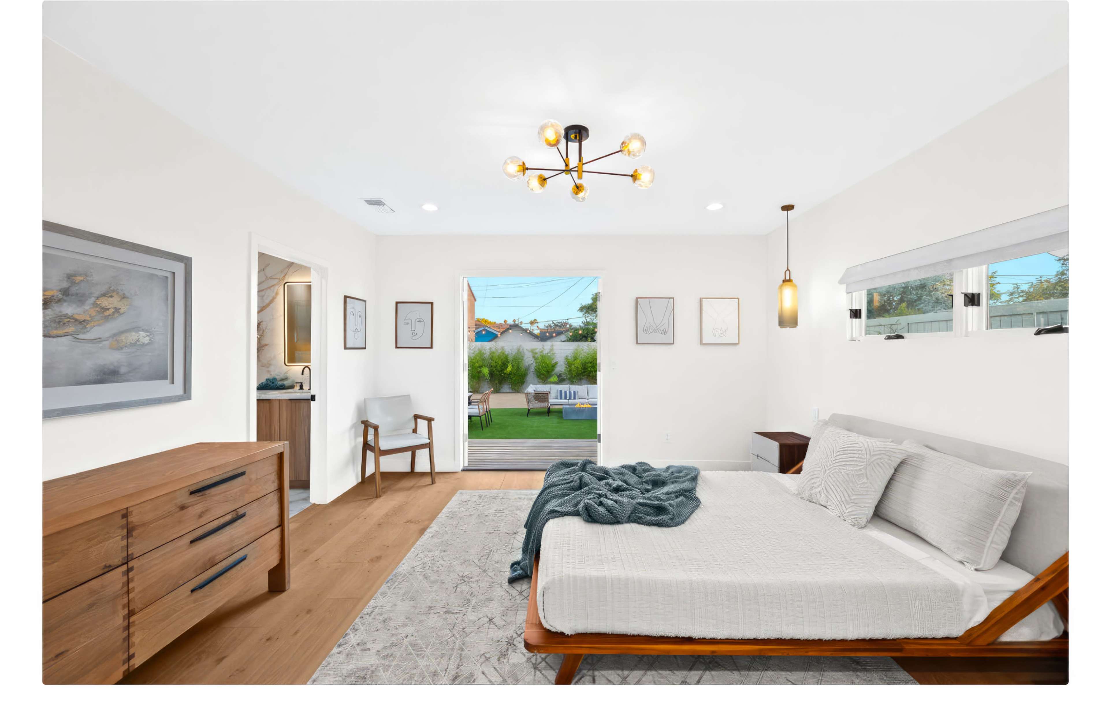 A modern bedroom features a bed without linens, a wooden dresser, and a view of a landscaped backyard through sliding doors.