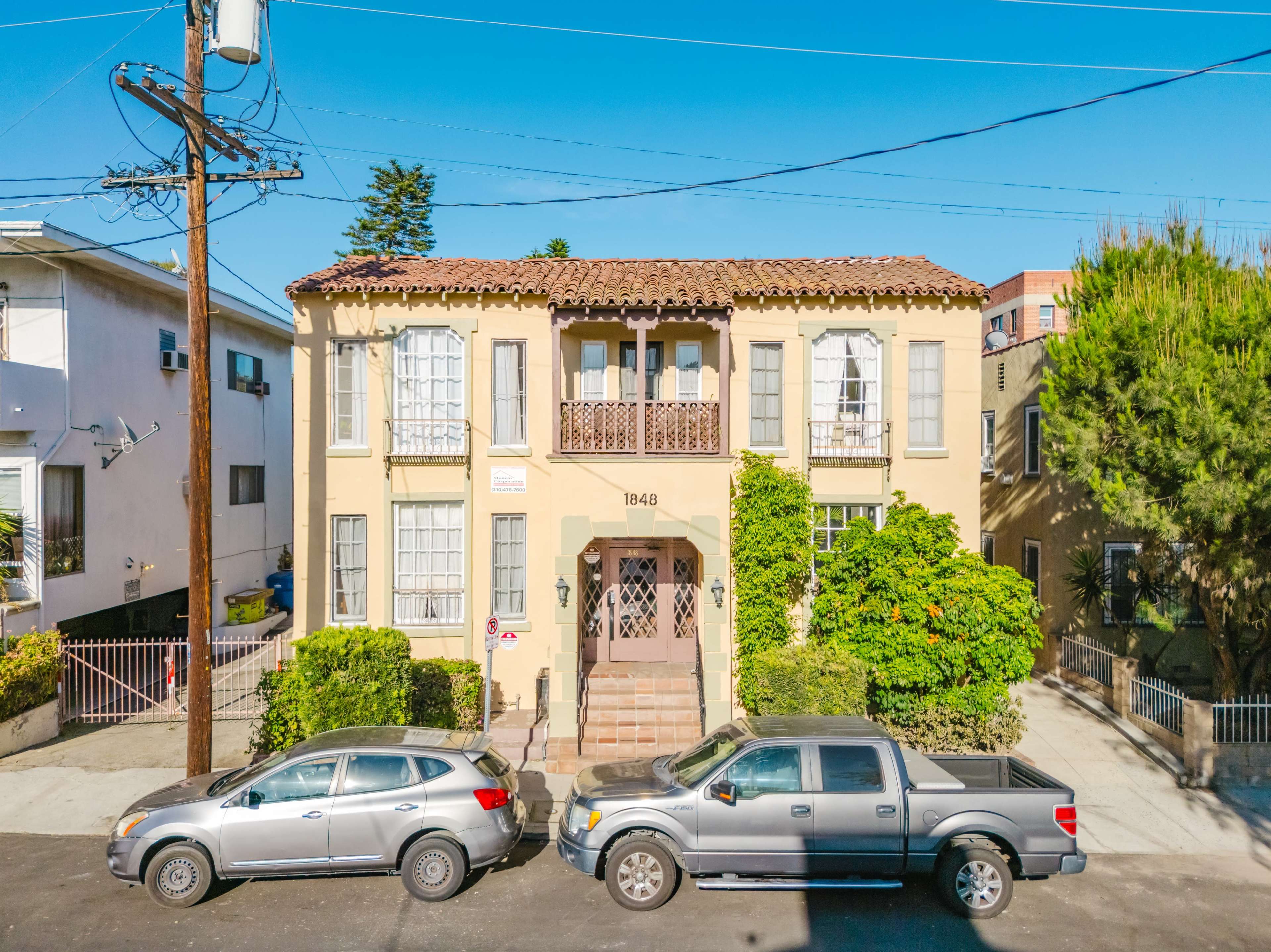 The image shows a two-story residential building with a stucco exterior, flanked by two parked vehicles on a quiet street.